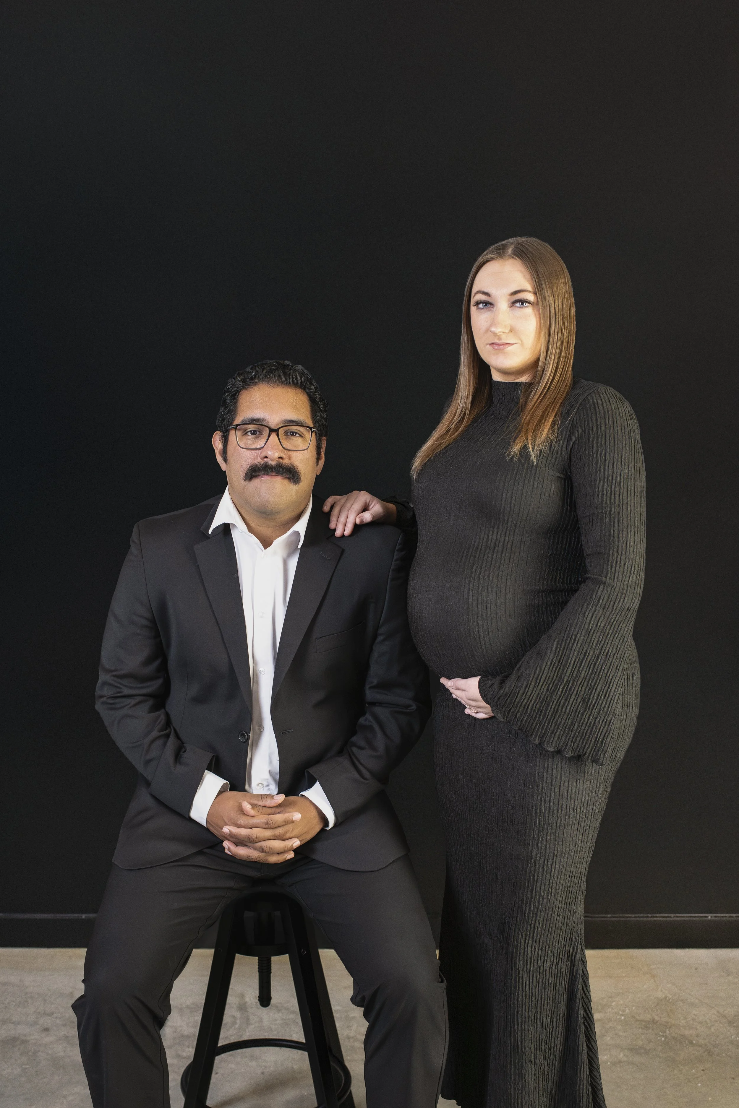 A man in a suit sitting on a stool with a woman in a black dress standing beside him against a dark background.