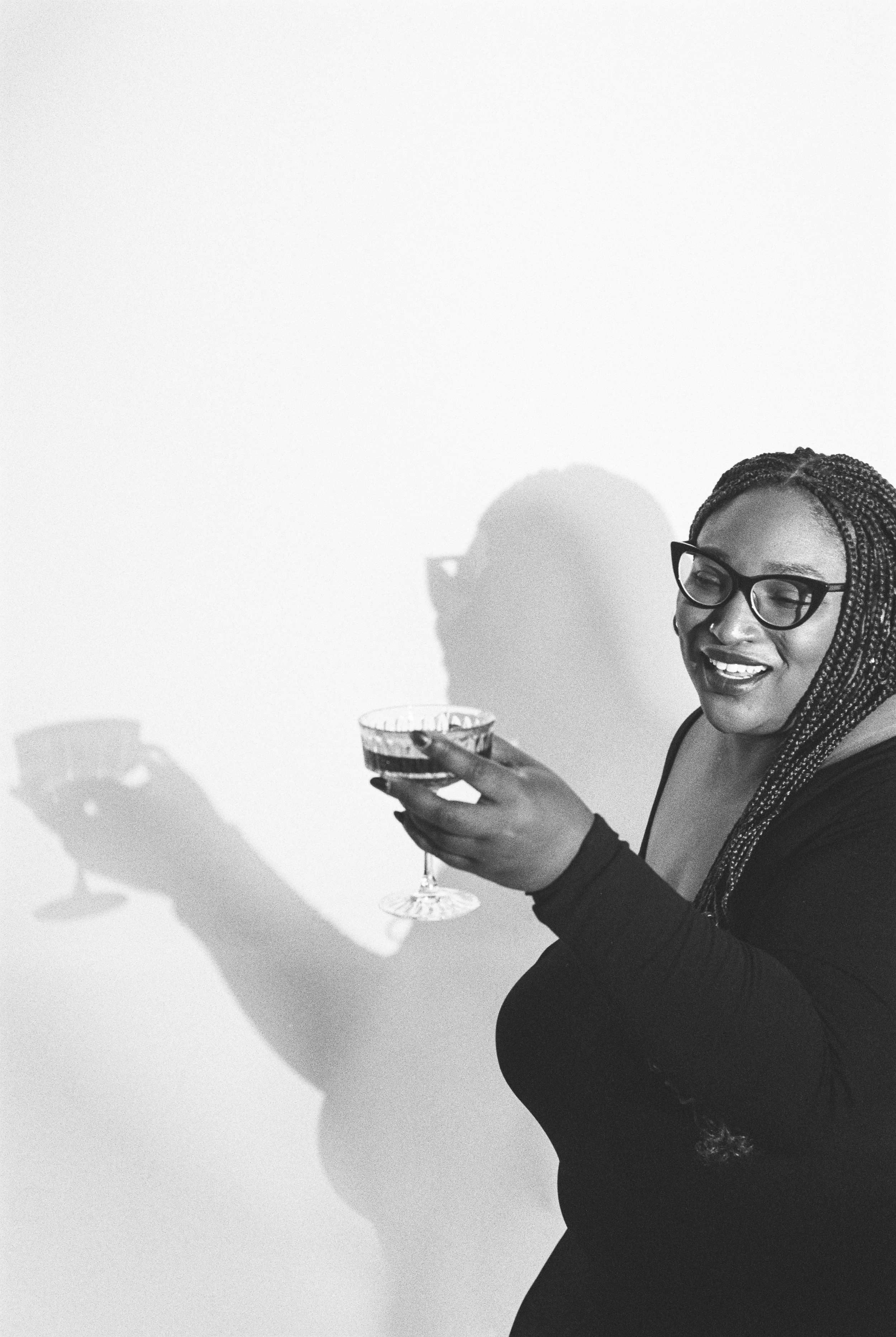 A woman with glasses and braids holding a glass of wine, smiling, with a shadow of herself holding a phone on the wall.