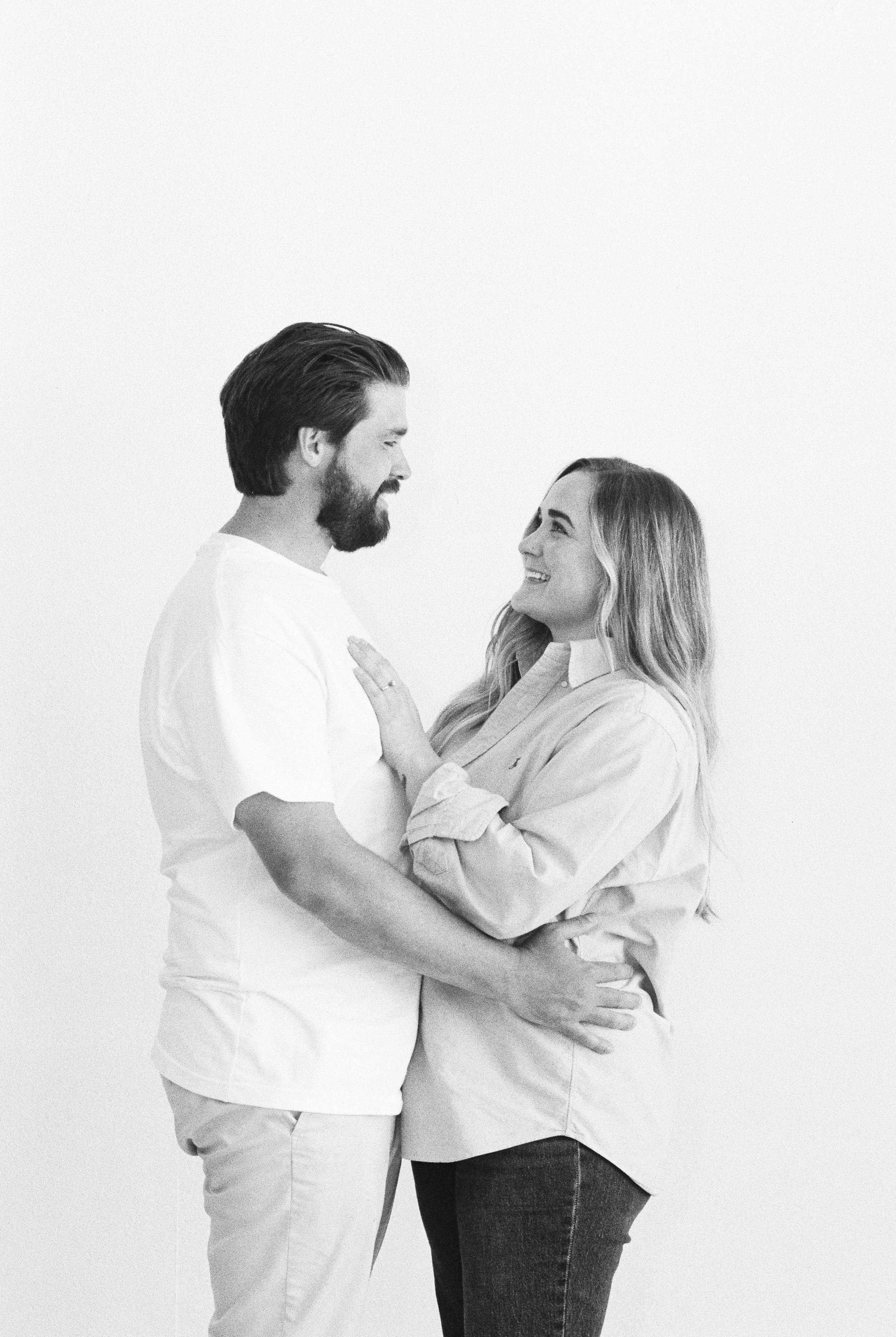 A black-and-white photo of a man and woman looking at each other affectionately, standing close together against a plain background.