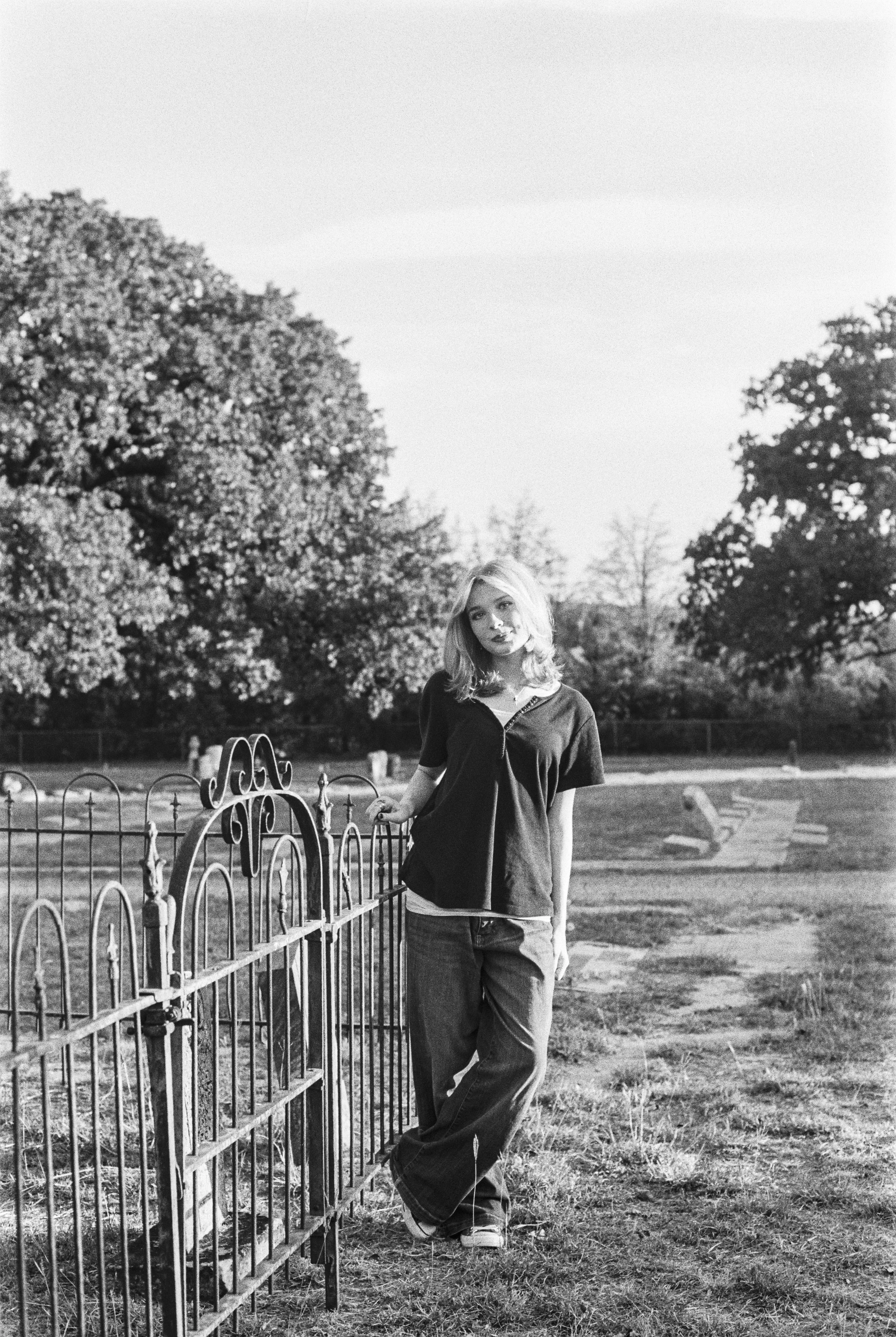 A young woman in casual clothing, standing beside a wrought iron fence, in a park with trees and benches in the background, captured in black and white.