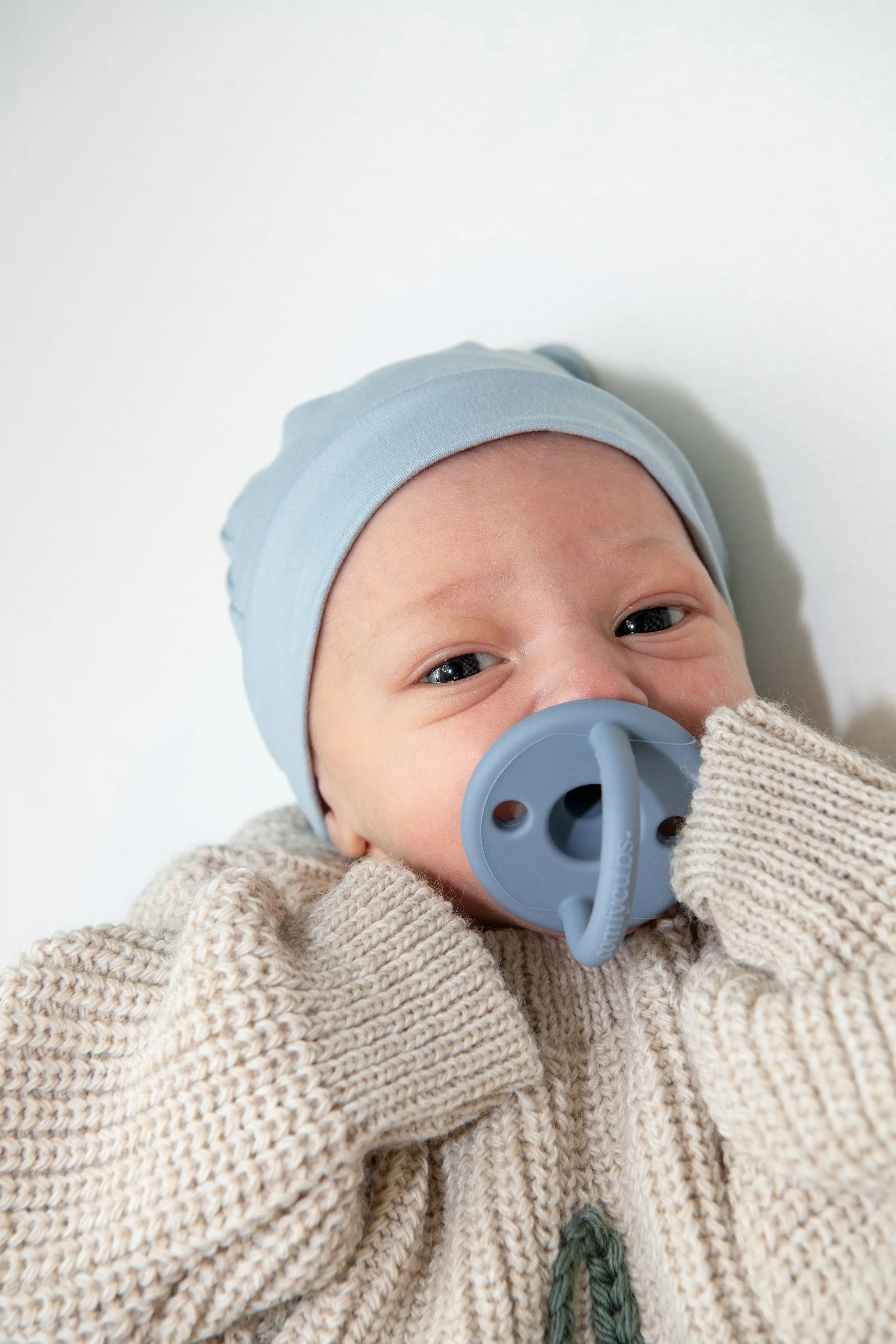 Close-up of a baby wearing a light blue hat and beige sweater, with a pacifier in their mouth, lying on a white surface.