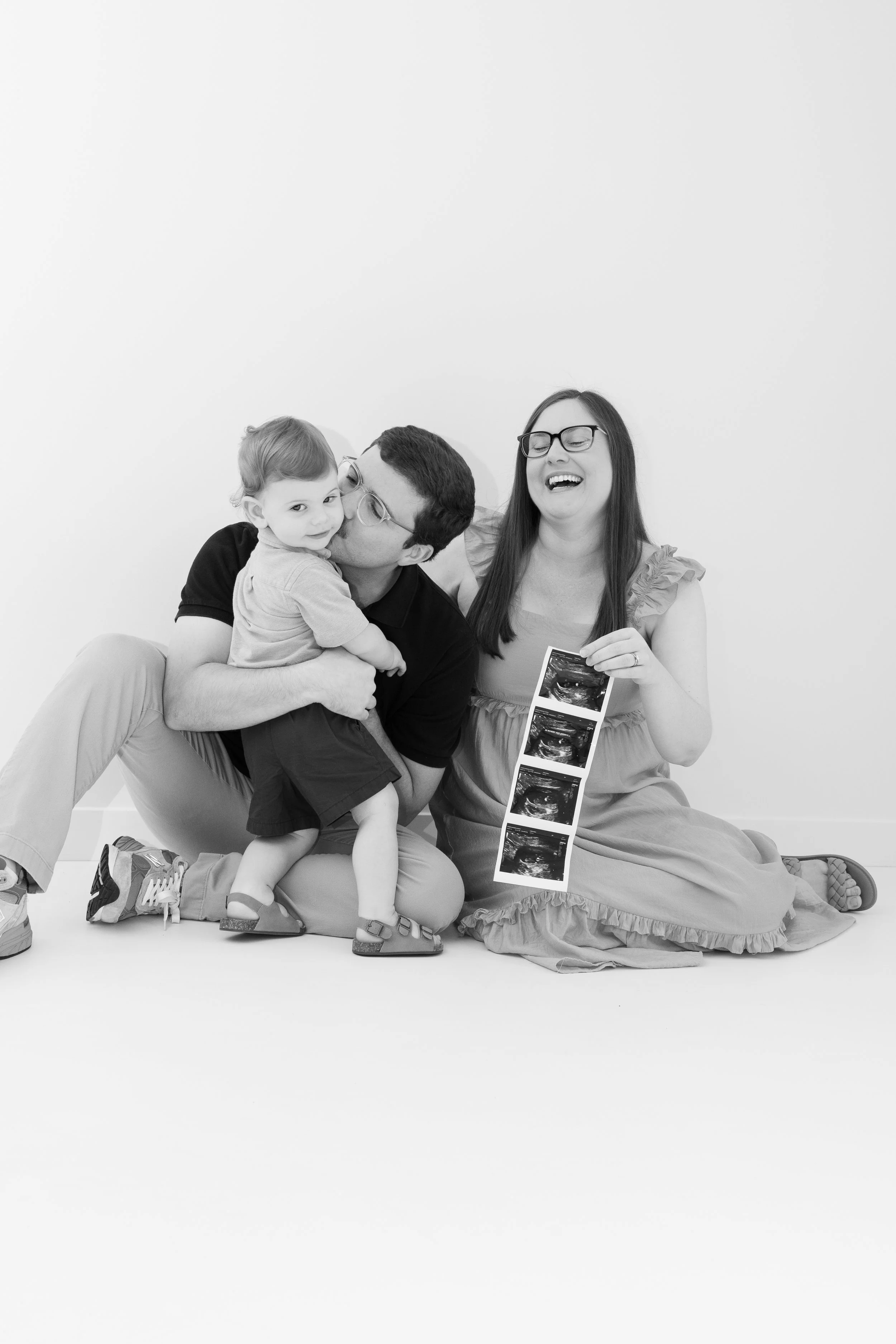 Black and white photo of a happy family, including a man, woman, and child, sitting on the floor with ultrasound photos, celebrating the pregnancy.