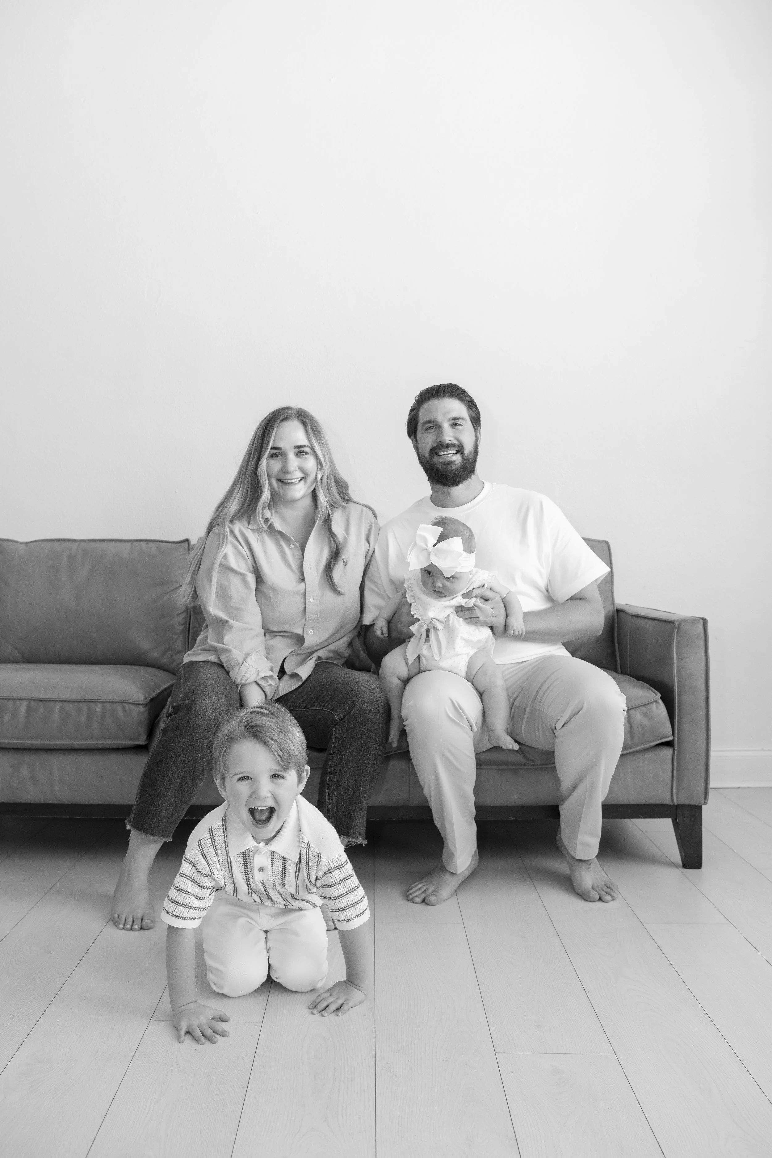 A black-and-white photo of a family of four sitting and kneeling on the floor in front of a sofa. The mother and father are sitting on the sofa, smiling at the camera, with a baby girl on the father's lap and a young boy kneeling on the floor, smilin