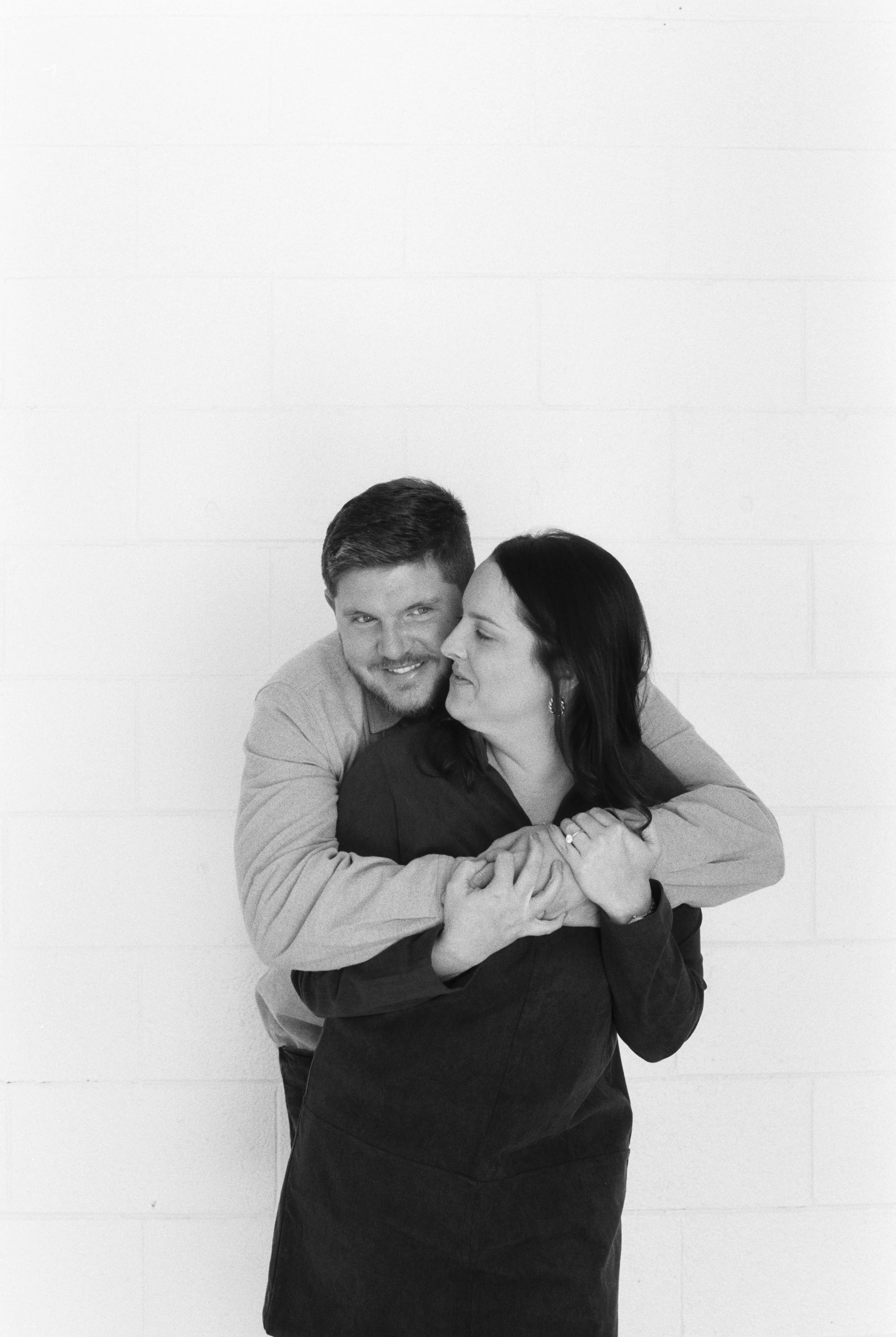 A black and white photo of a couple hugging and smiling, standing against a plain white wall.