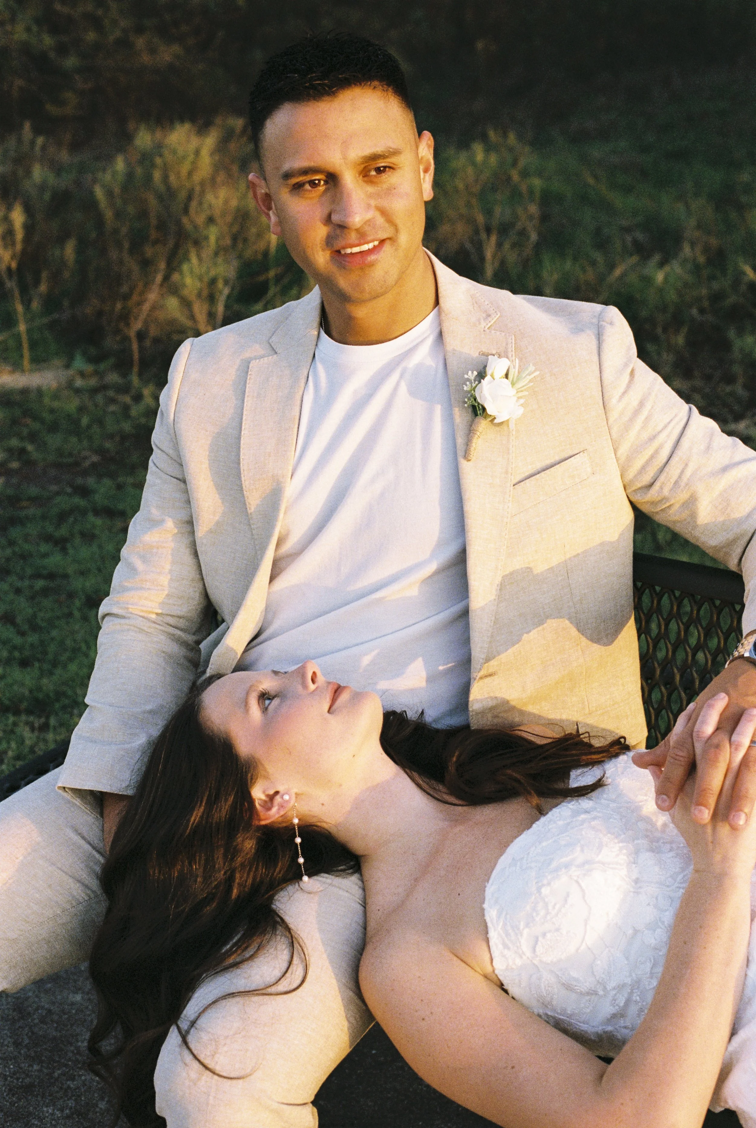 A man and woman sitting on a bench outdoors during sunset. The man is wearing a light-colored suit with a white shirt and has a boutonniere on his lapel. The woman, lying on the man's lap, is wearing a white strapless dress, looking up at him, holdin