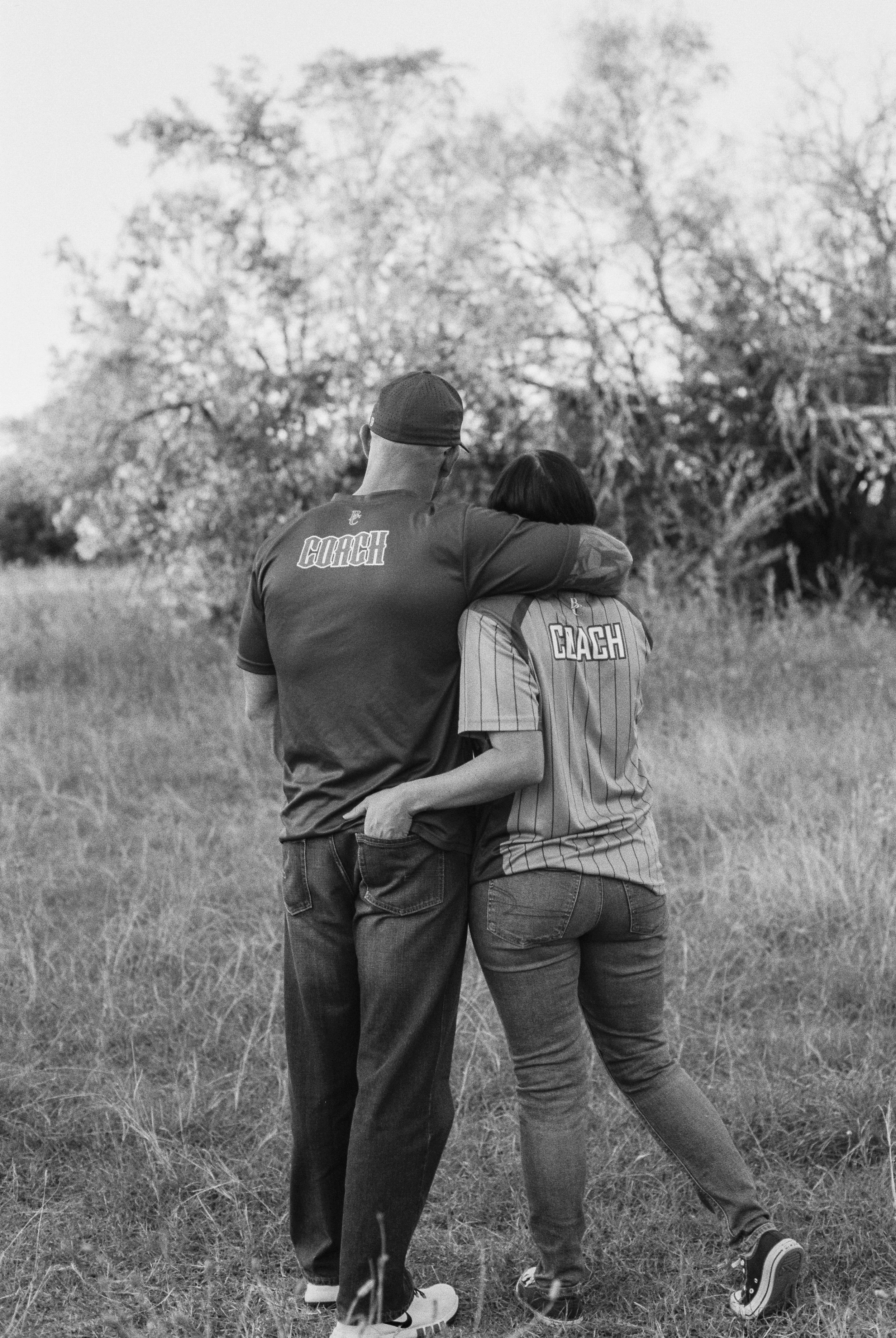 A black and white photo of a man and woman walking in a grassy field, with the man’s arm around the woman’s shoulders. Both wear shirts labeled 'COACH' on the back, and the woman leans into the man as they walk away from the camera.