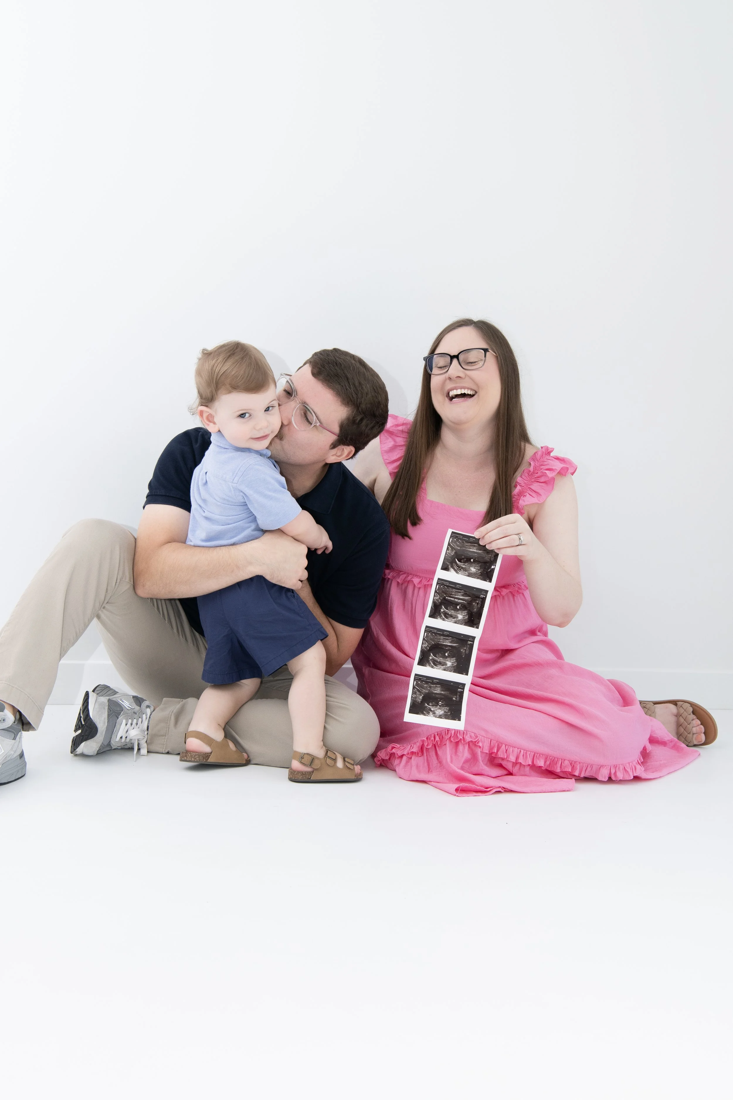 A family of three sitting on the floor, celebrating a pregnancy with an ultrasound printout. The mother, wearing a pink dress, is holding ultrasound images and laughing. The father, in a navy shirt and beige pants, is kissing a young girl who is sitt