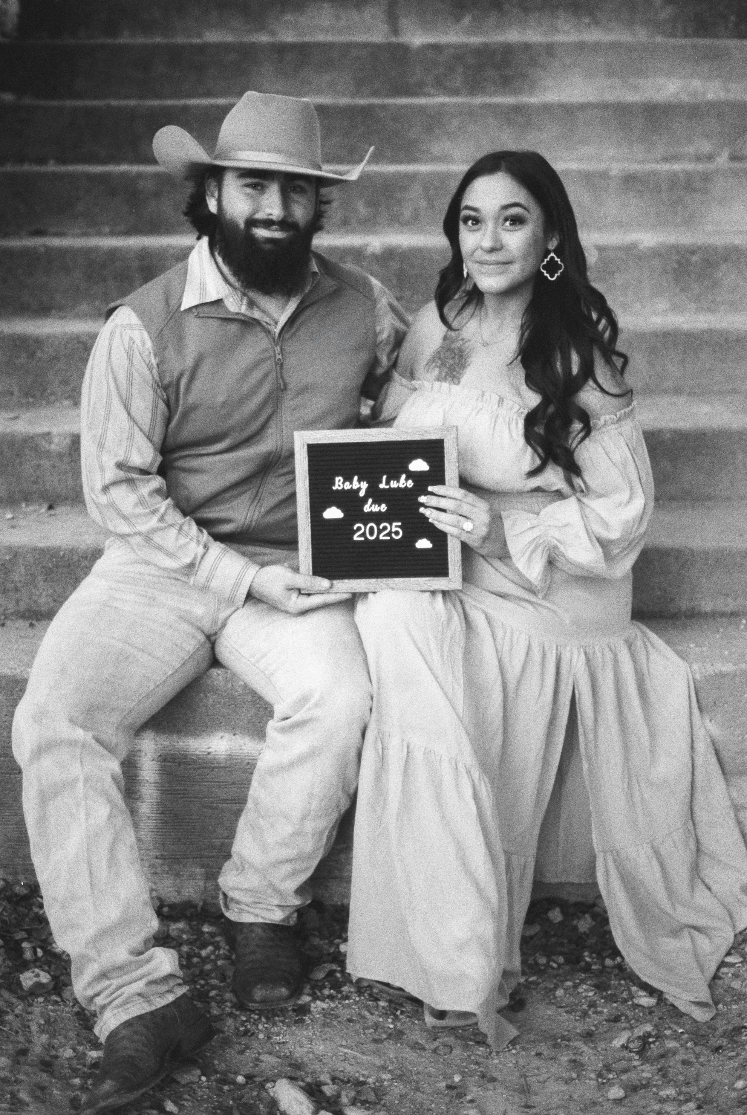 A couple sitting on outdoor stairs holding a sign that reads 'Baby Luke due 2025'.