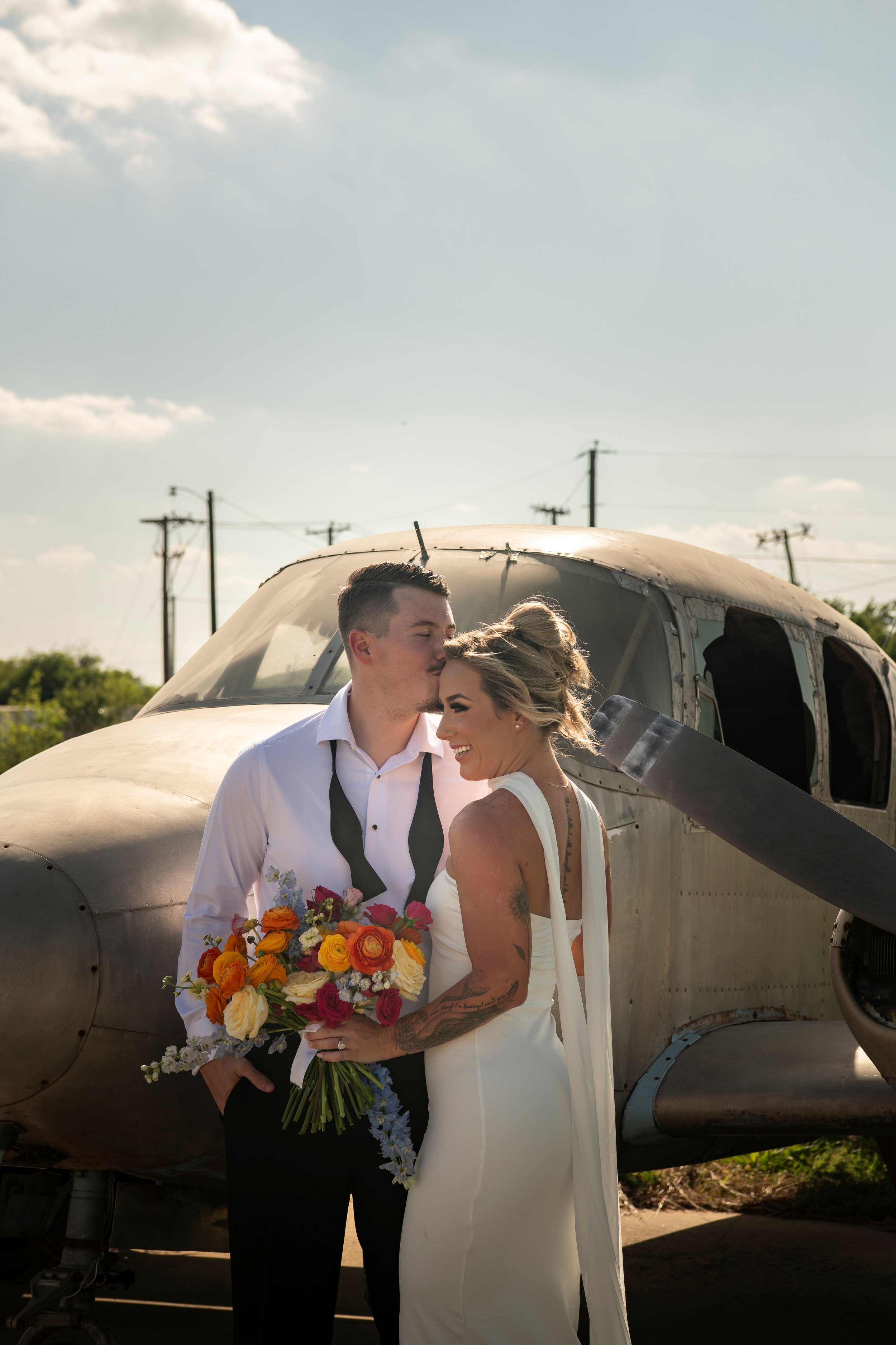 A newlywed couple standing in front of a vintage airplane. The groom is holding a colorful bouquet and kissing the bride's forehead. The bride is smiling, wearing a white dress with black tattoos visible on her arms. The airplane has a metal body and