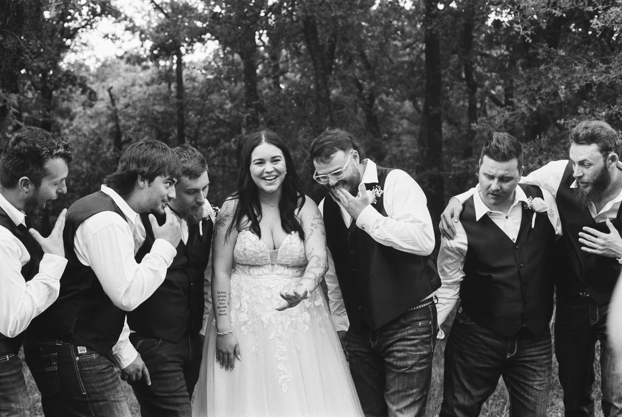 Black and white photo of a bride surrounded by groomsmen in an outdoor setting with trees in the background.