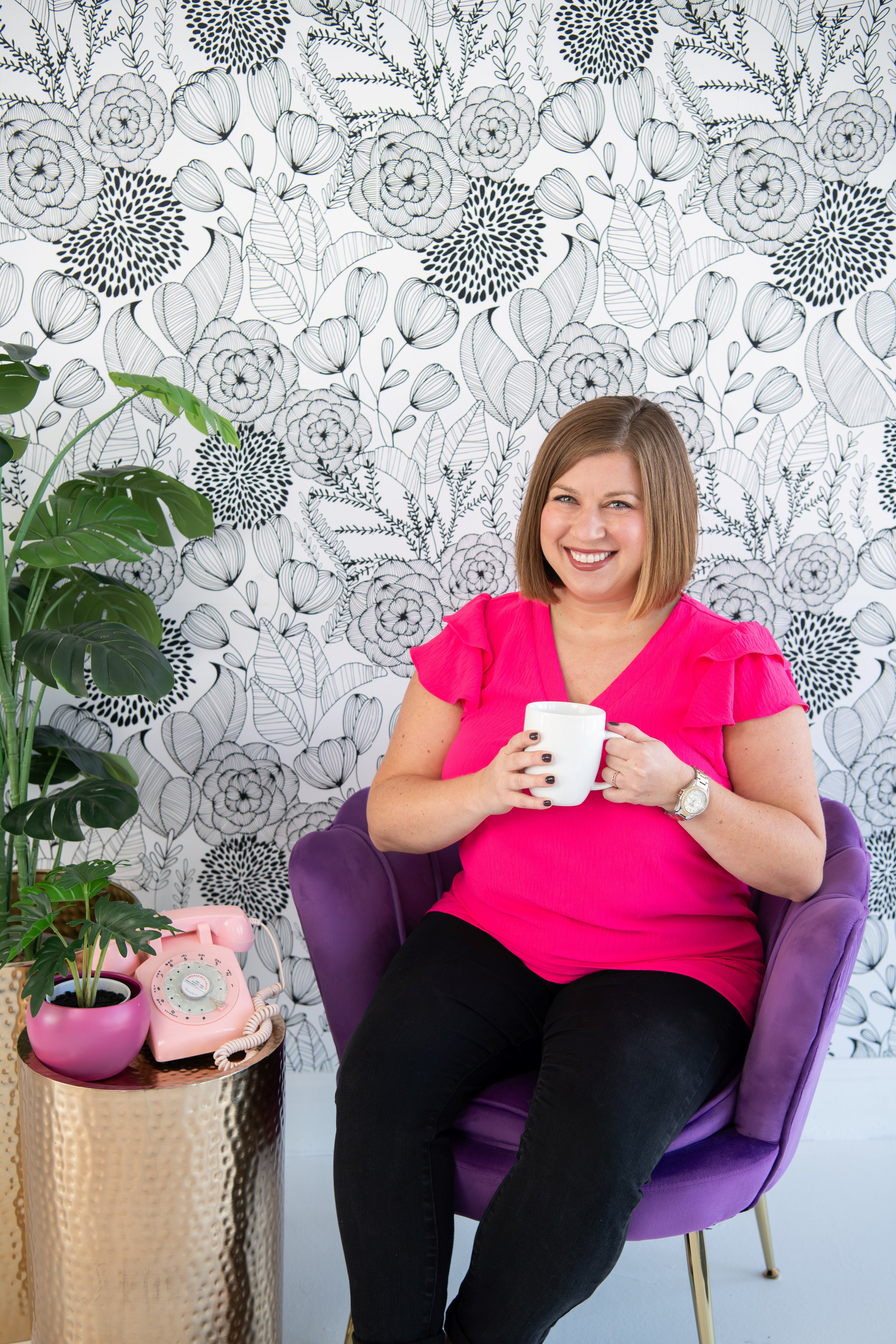 A woman with shoulder-length hair, wearing a bright pink top, is sitting in a purple armchair, smiling and holding a white mug. Behind her is a black and white floral patterned wallpaper. To her left, there is a small round gold table with a pink vintage rotary phone, a pink pot with a green plant, and a gold vase.