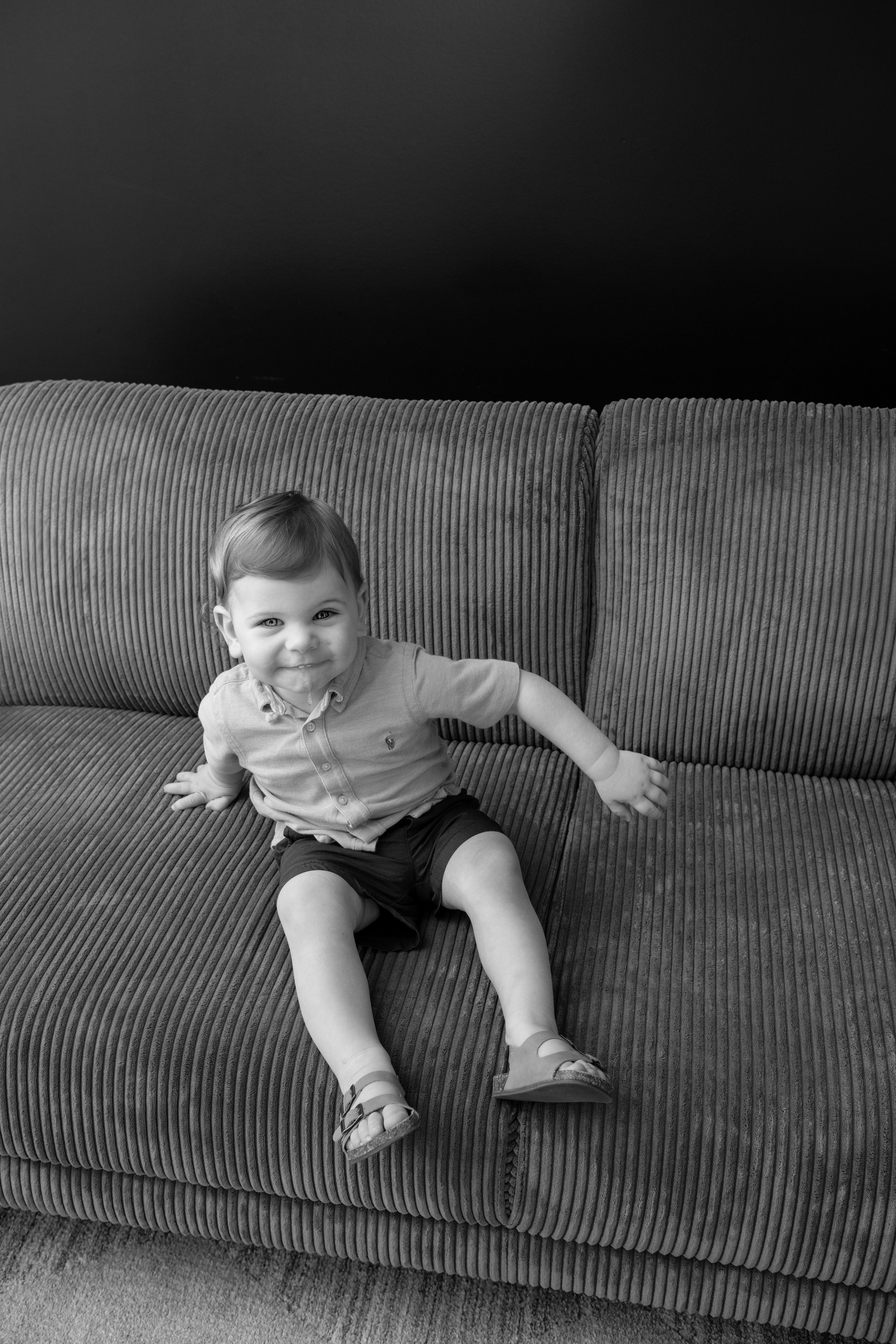 A smiling young child sitting on a corduroy sofa with a dark wall in the background.