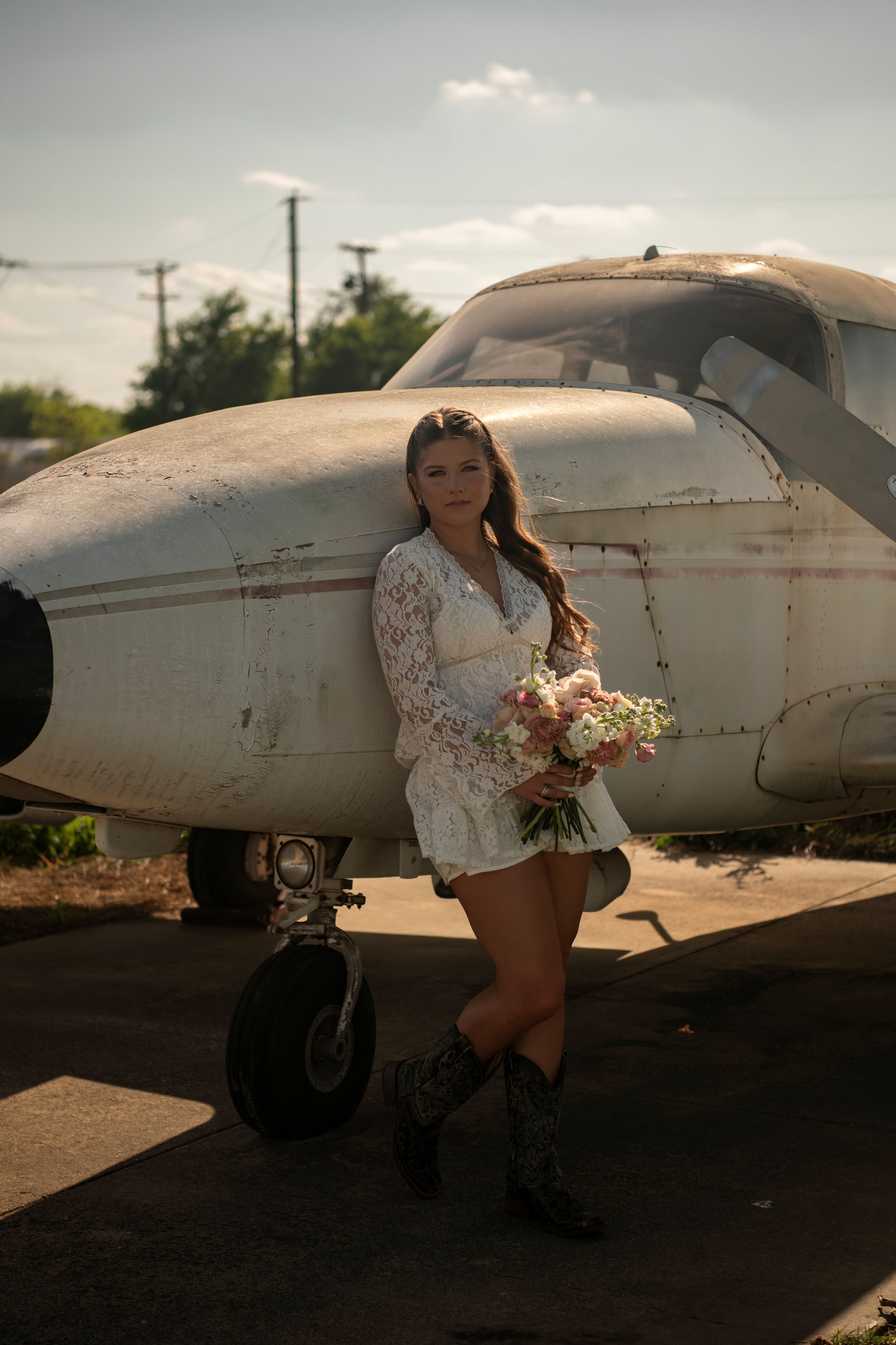 A woman wearing a white lace dress holding a bouquet of flowers posing in front of a small, vintage airplane. The scene is outdoors during daytime with a partly cloudy sky and utility poles in the background.