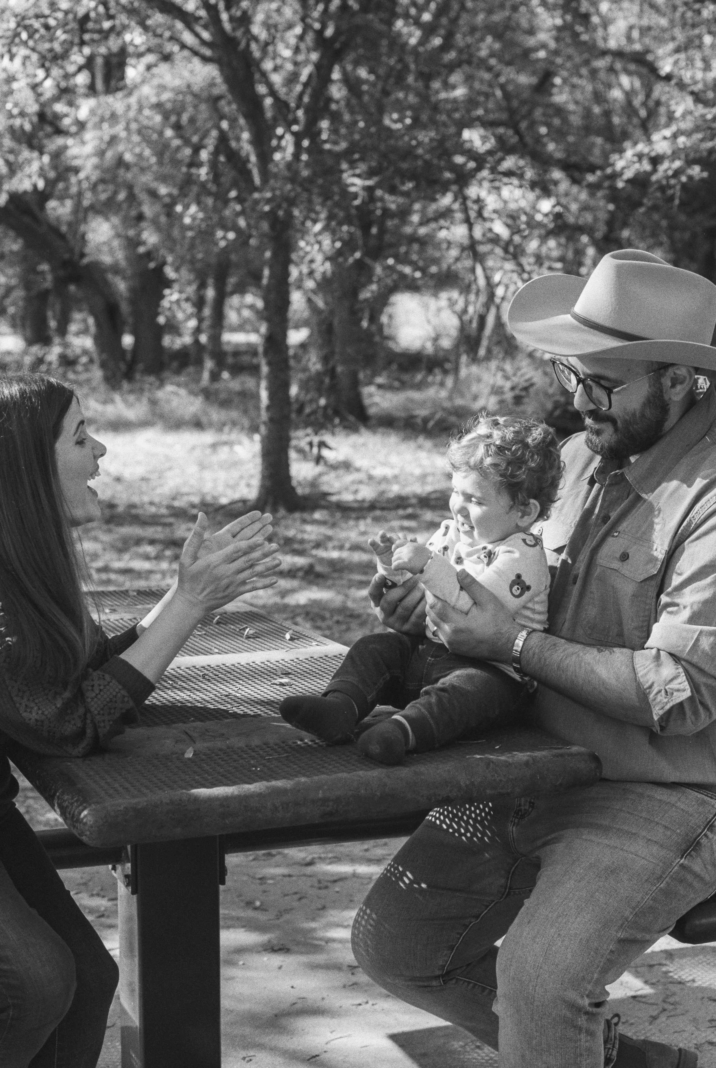 A black and white photo of a family outdoors, with a woman and a man playing with a young child on a picnic table in a park with trees in the background.