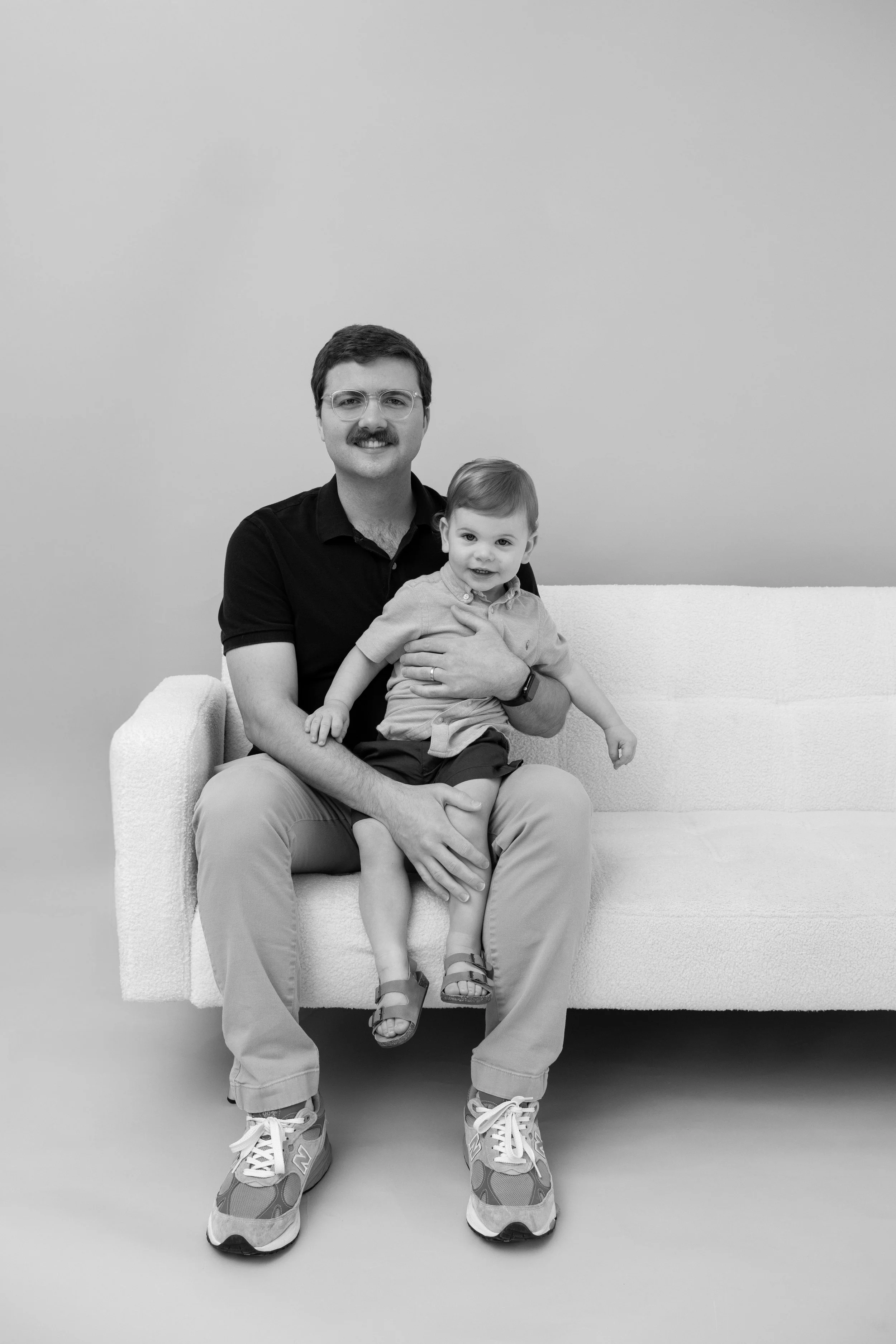 A man with glasses and a mustache sitting on a sofa, holding a young boy on his lap, both smiling at the camera in a black and white photo.