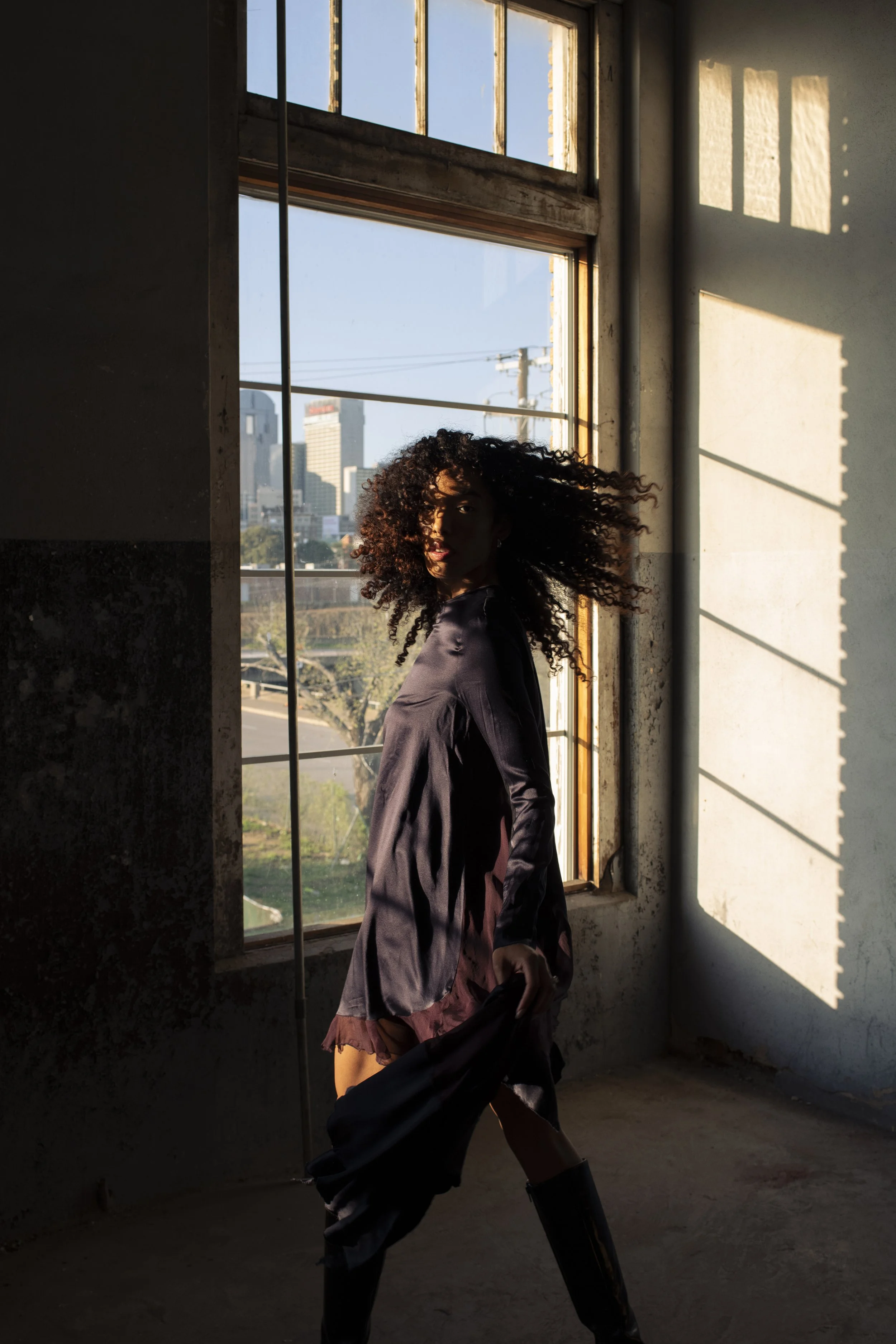 A woman with curly hair and a dark jacket standing inside a building near a large window. The sunlight coming through the window casts shadows on the wall. The city skyline with tall buildings and a clear blue sky is visible outside.