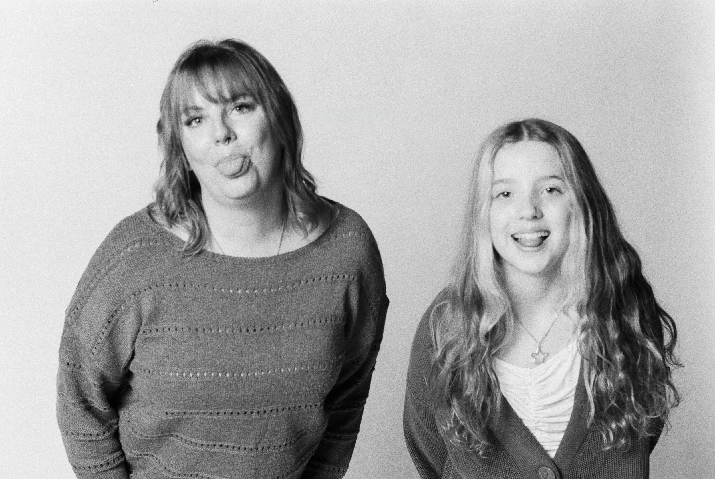 Black and white photo of a woman and a girl standing side by side against a plain background, both sticking out their tongues and smiling.