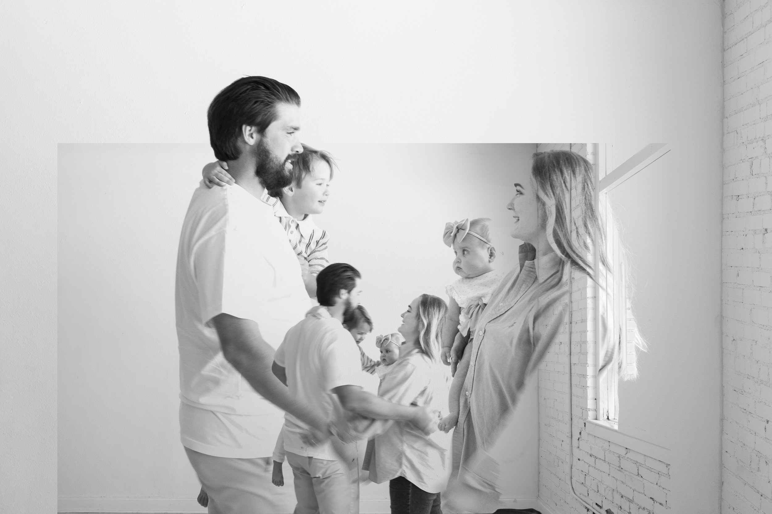 A black and white photo of a family group holding hands and smiling together, facing each other, in a bright room with a window and brick wall.