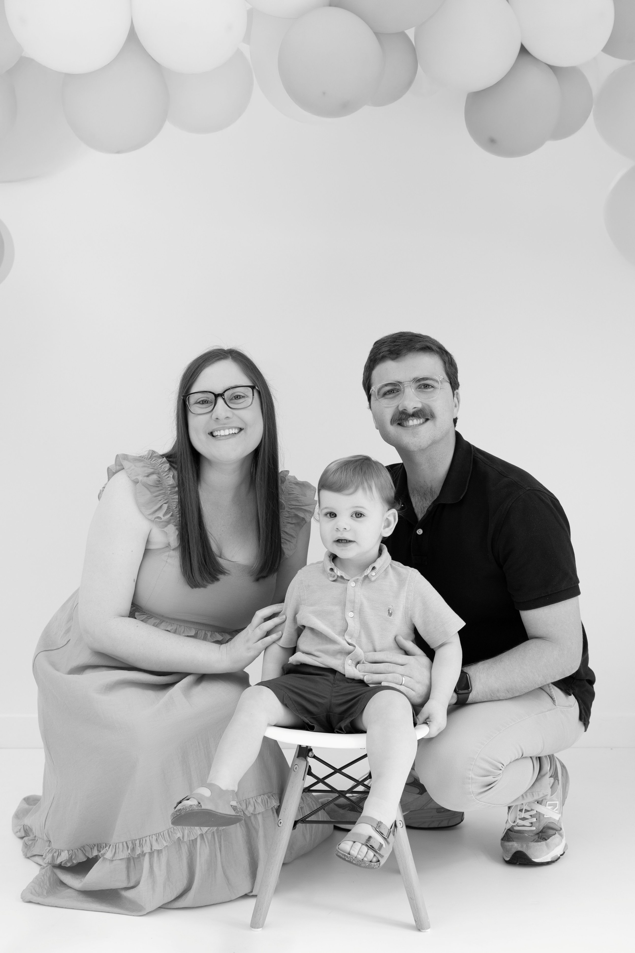 Black and white photo of a smiling family of three, a woman with straight hair and glasses, a man with mustache and glasses, and a young boy sitting on a small chair in front of them, with balloons hanging from the ceiling above.