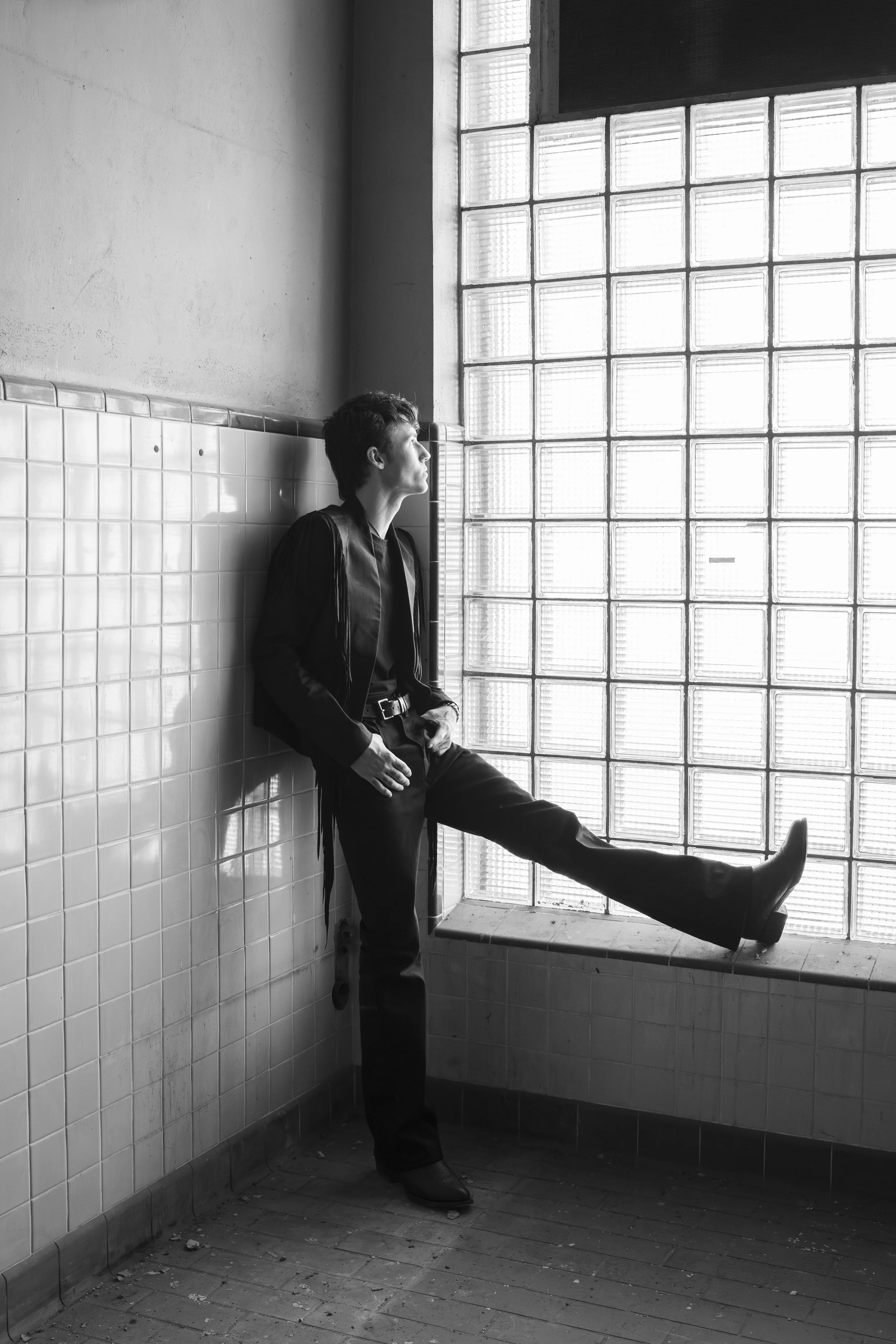 A young man stands in a corner of a room with tiled walls and a large glass block window, leaning against the wall with one hand in his pocket and his right leg extended onto the window ledge, dressed in dark clothing and wearing boots.