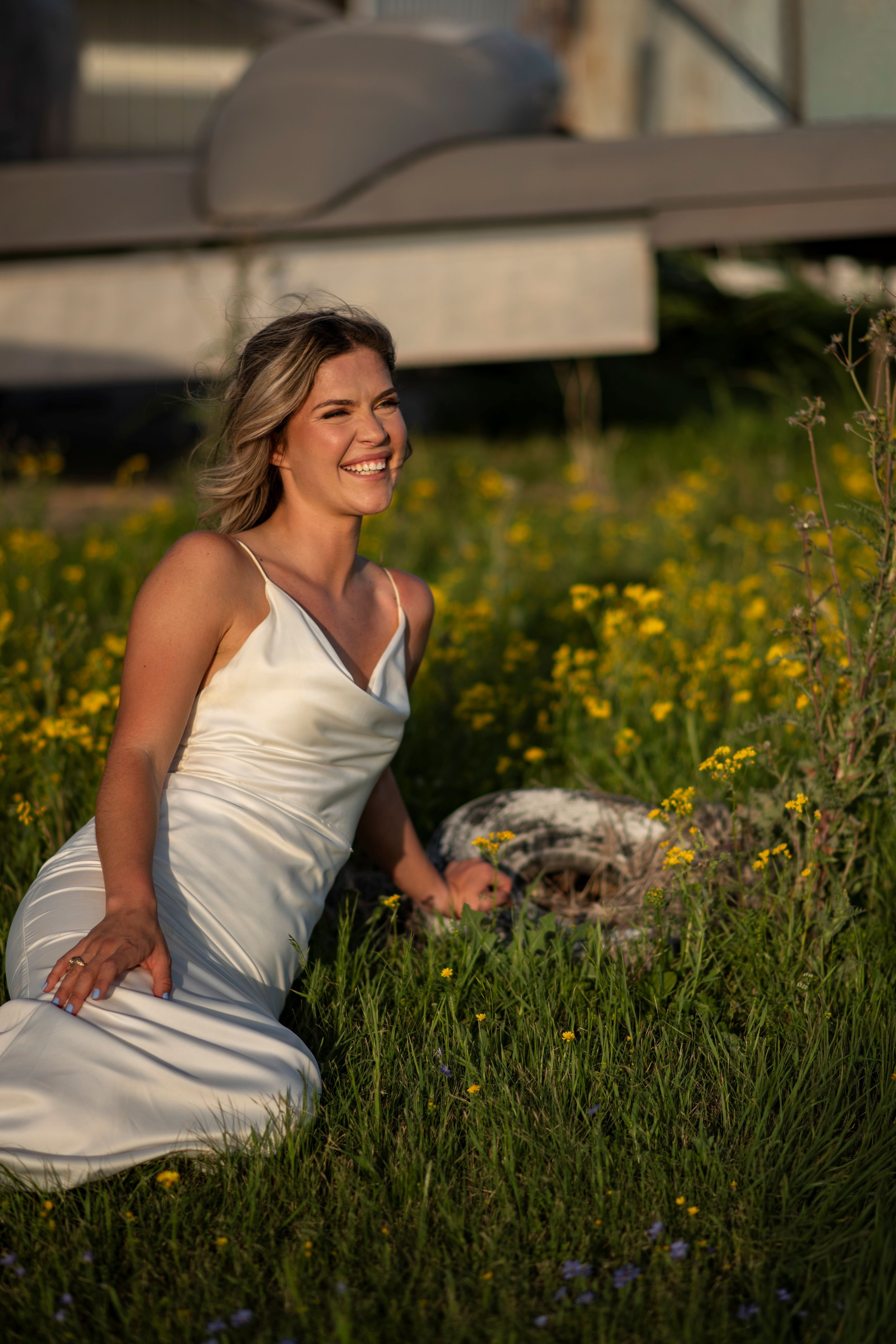 A woman in a white dress sitting on grass in a field of yellow flowers, smiling as she leans on her left hand, with an overpass or bridge above her.