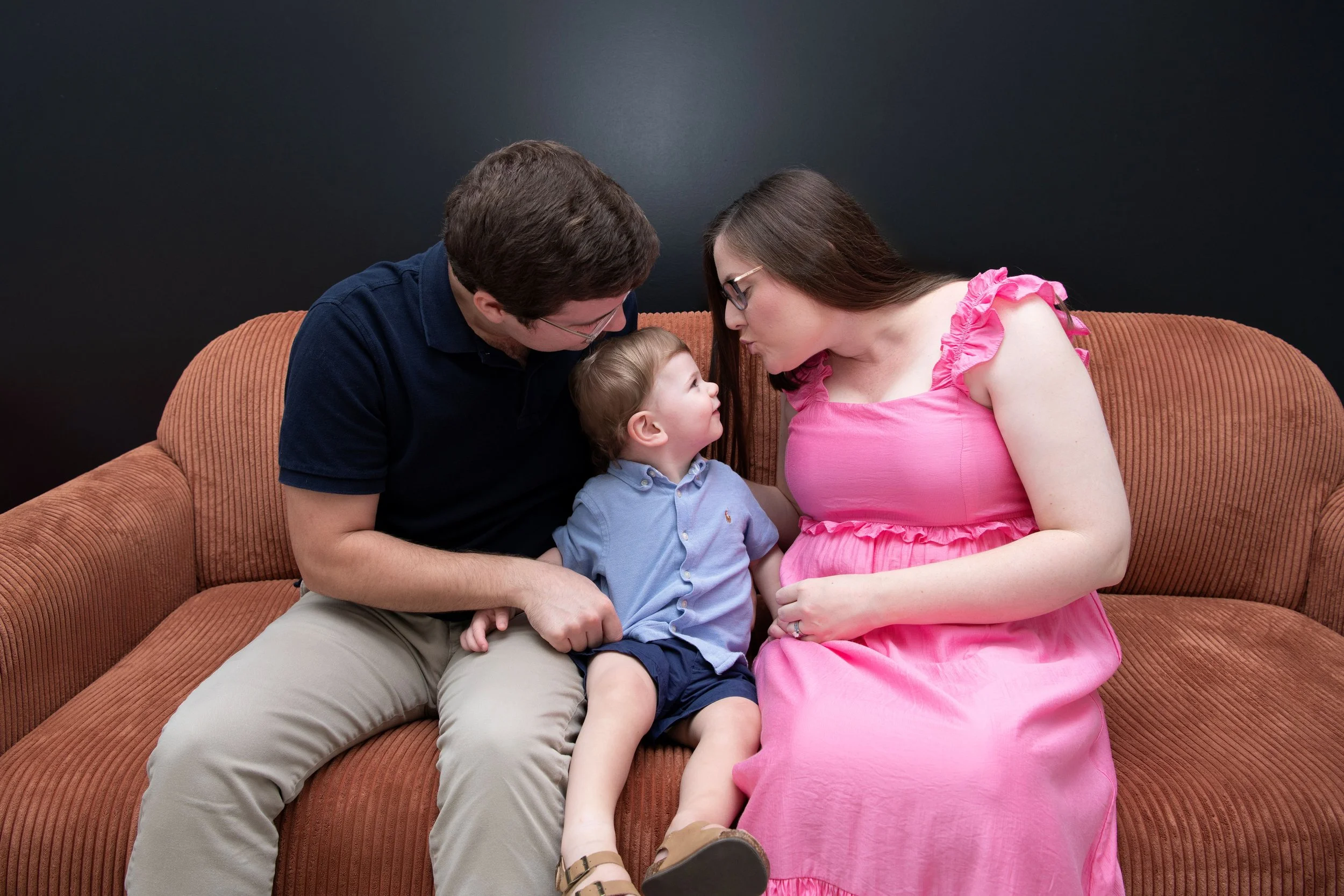A family of three sitting on a brown couch against a black background, with a woman in a pink dress, a young boy in a blue shirt, and a man in a black shirt.