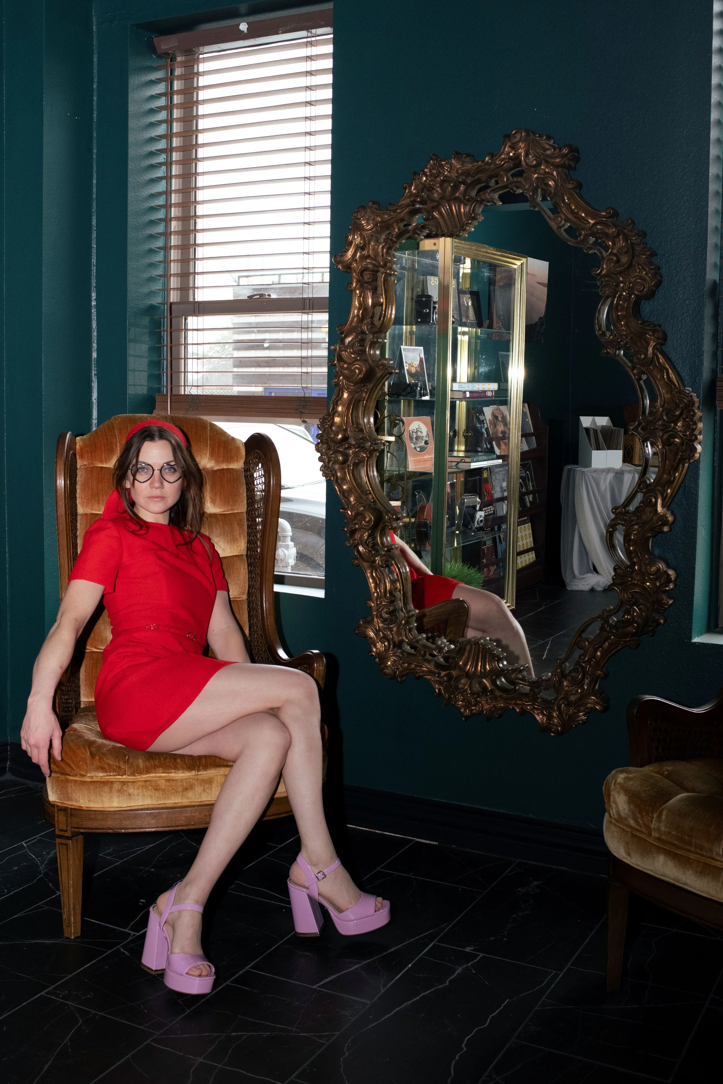 A woman in a red dress sitting on a vintage armchair in a room with teal walls, a window, and a large ornate mirror reflecting her image.