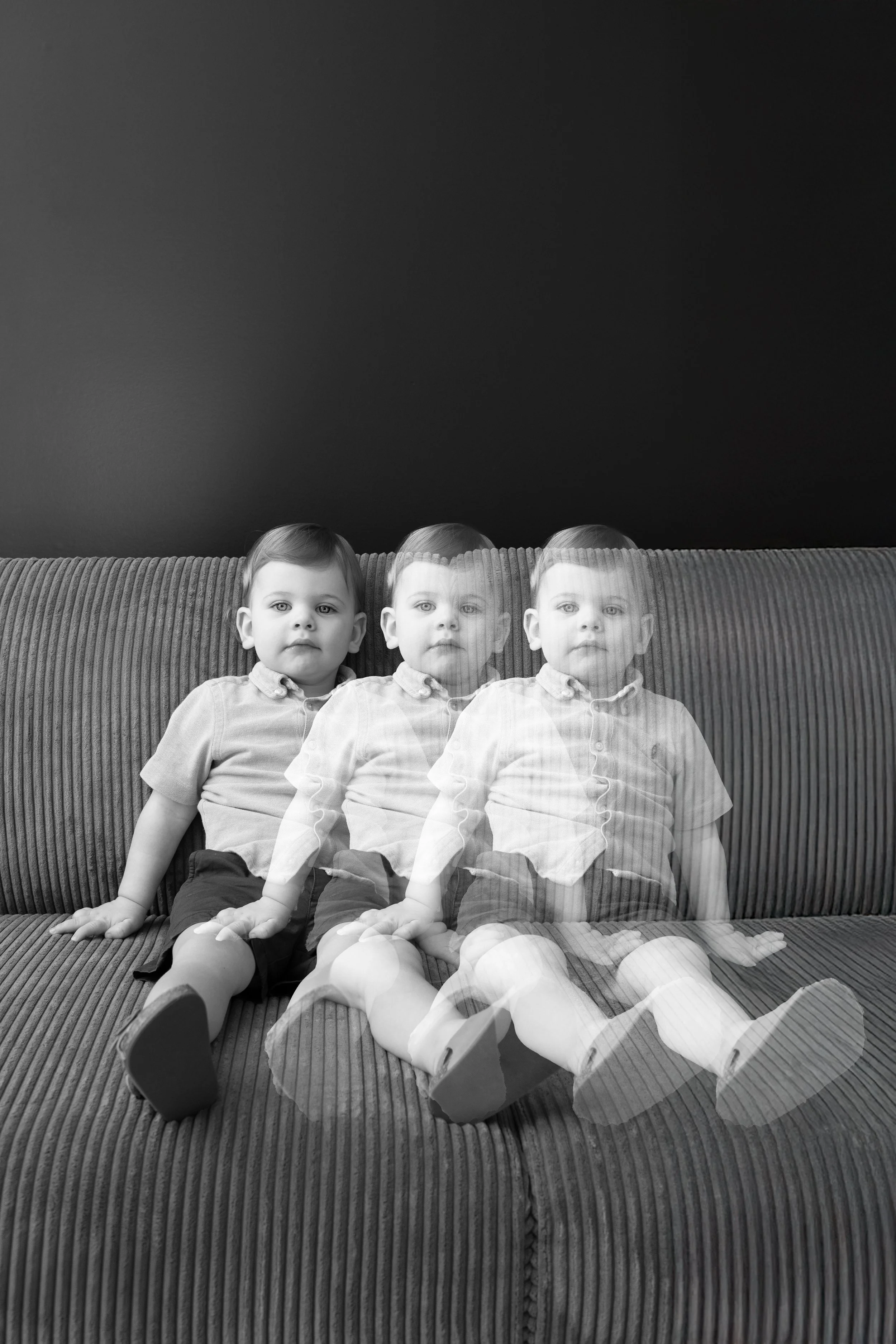 A black and white photograph of a young boy sitting on a textured sofa against a plain dark wall, with a three-exposure effect showing him in three different positions.