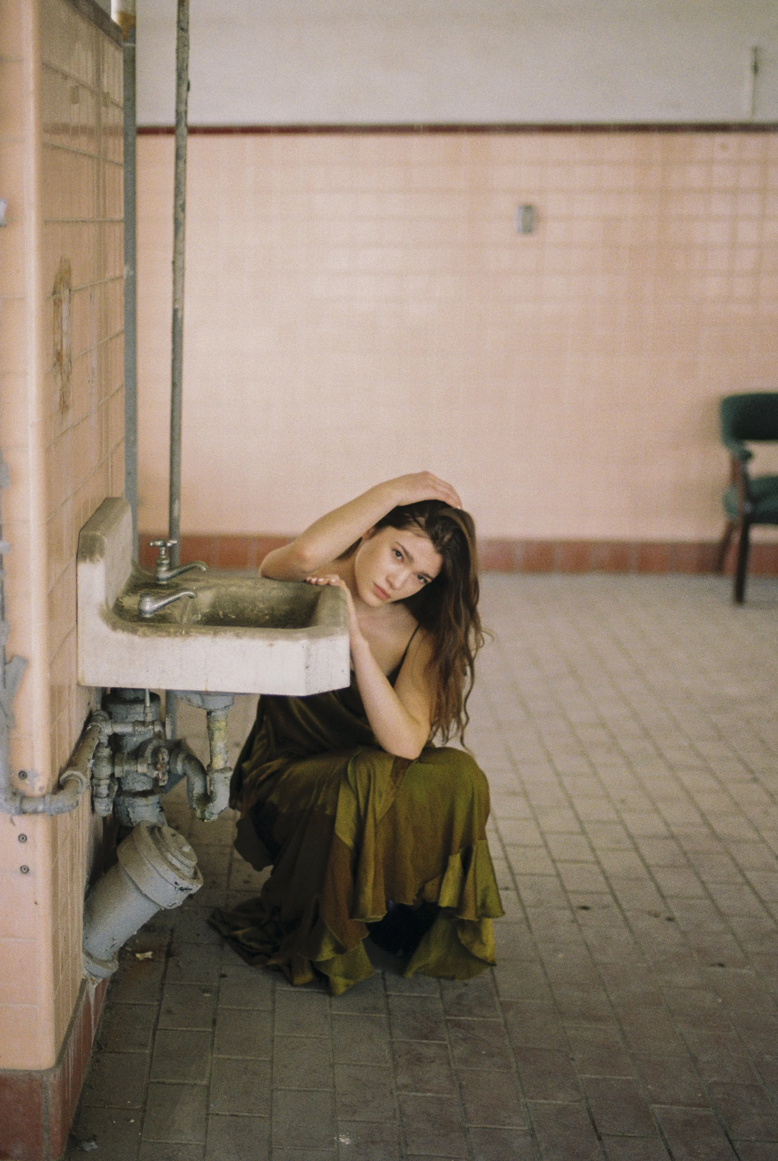 A woman with long brown hair crouches near a wall and sink in a dimly lit, industrial-style room. She wears a dark top and a flowing, yellow-brown skirt. She rests her head and hands on her left arm, looking directly at the camera.