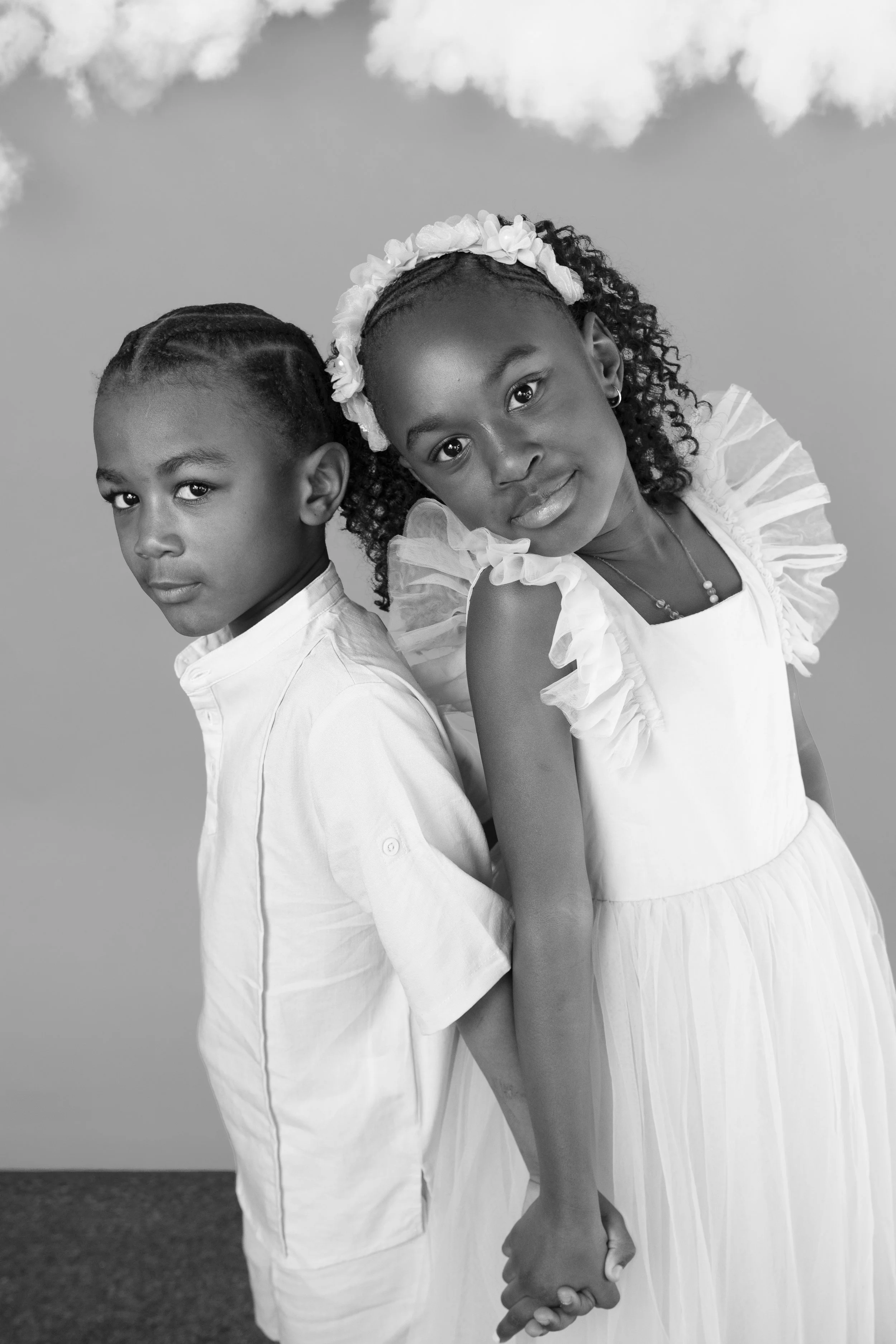 Two children, a girl and a boy, standing back-to-back against a cloudy sky background. The girl is wearing a white dress with ruffled sleeves and a floral headband, and is holding the boy's hand. Both have serious expressions, with the girl slightly 