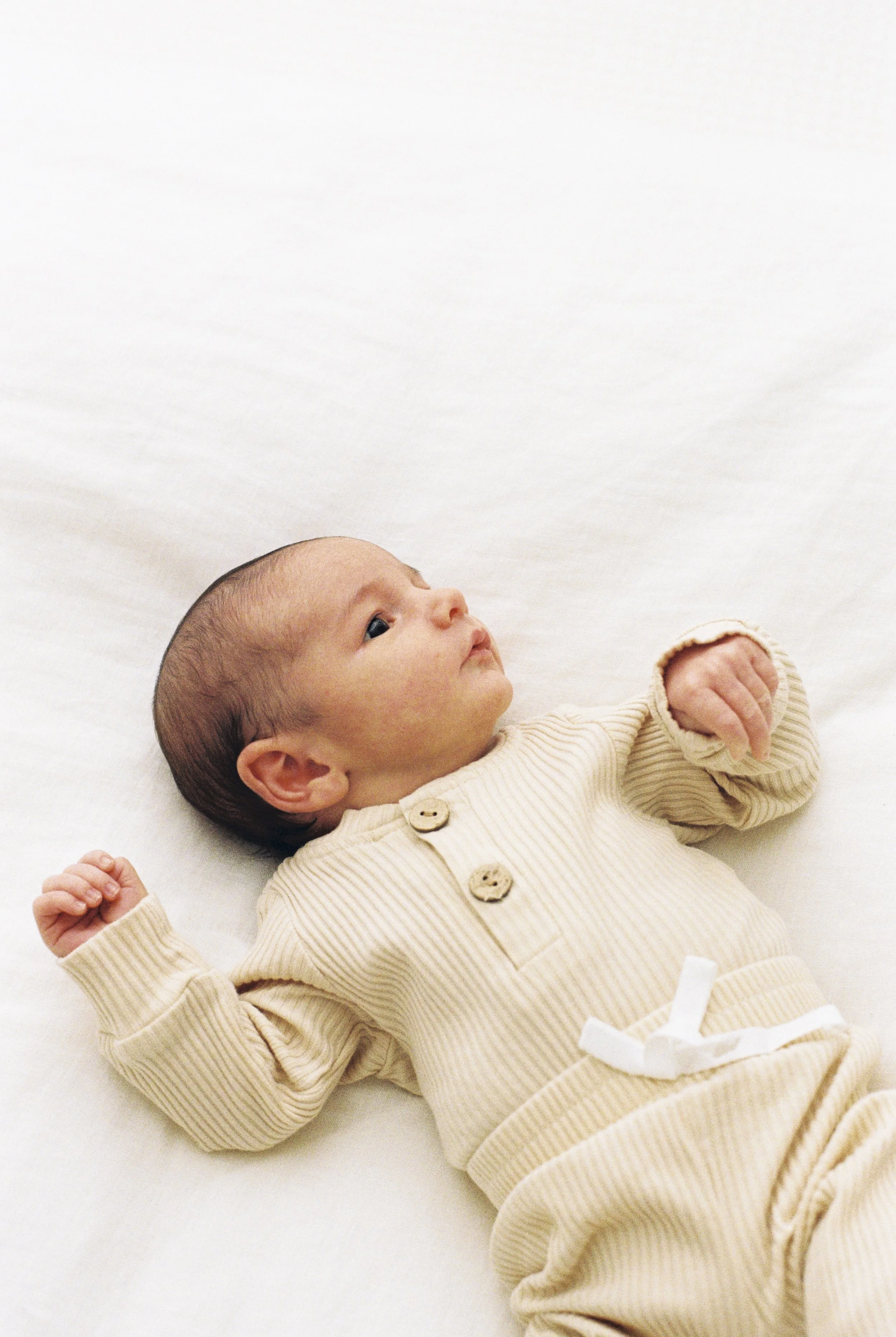 A baby lying on a white surface, looking upwards with arms slightly raised, wearing a beige ribbed outfit with buttons and a white bow at the waist.