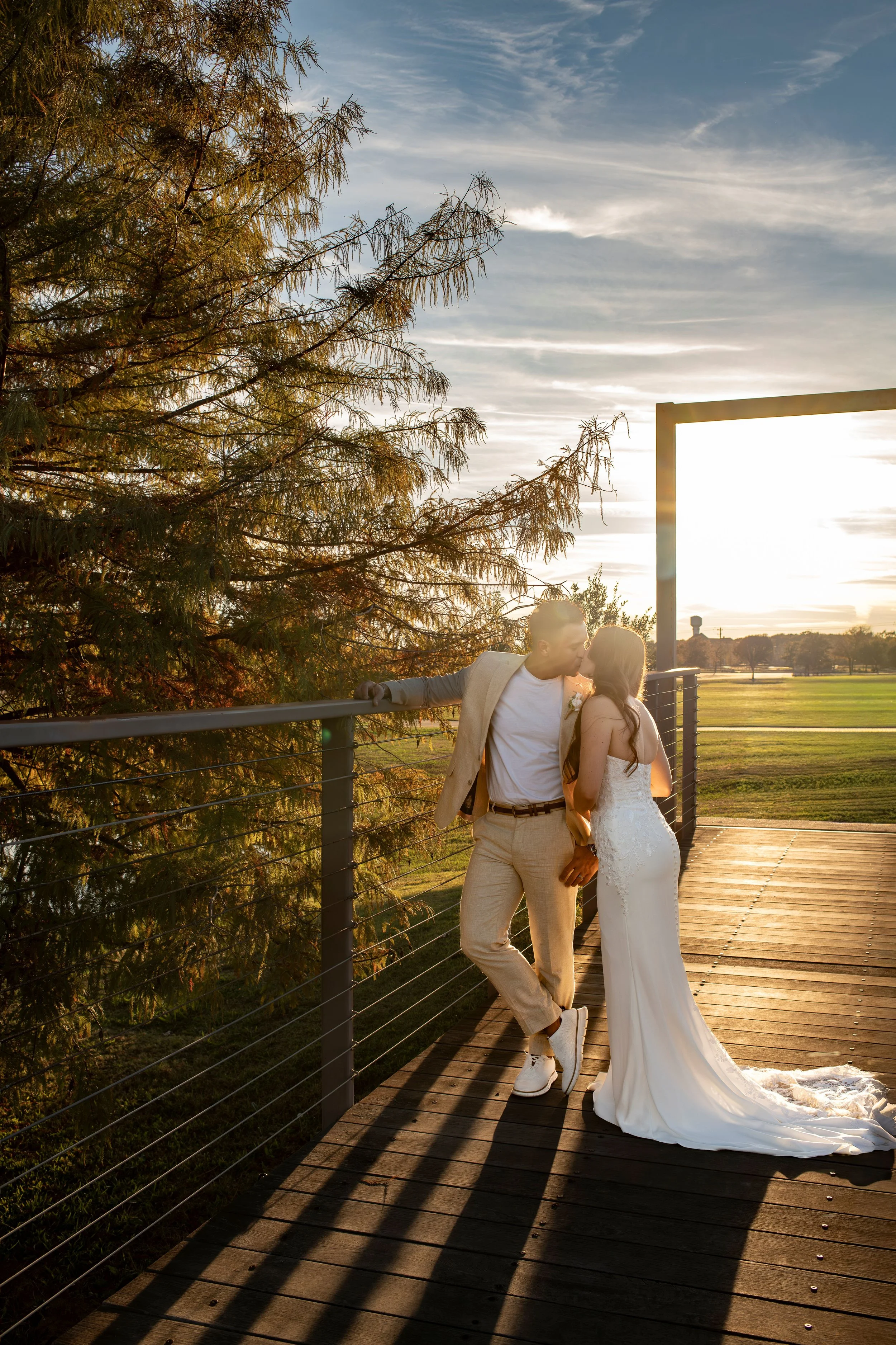 A couple kissing on a wooden deck at sunset, with a large tree and open field in the background.