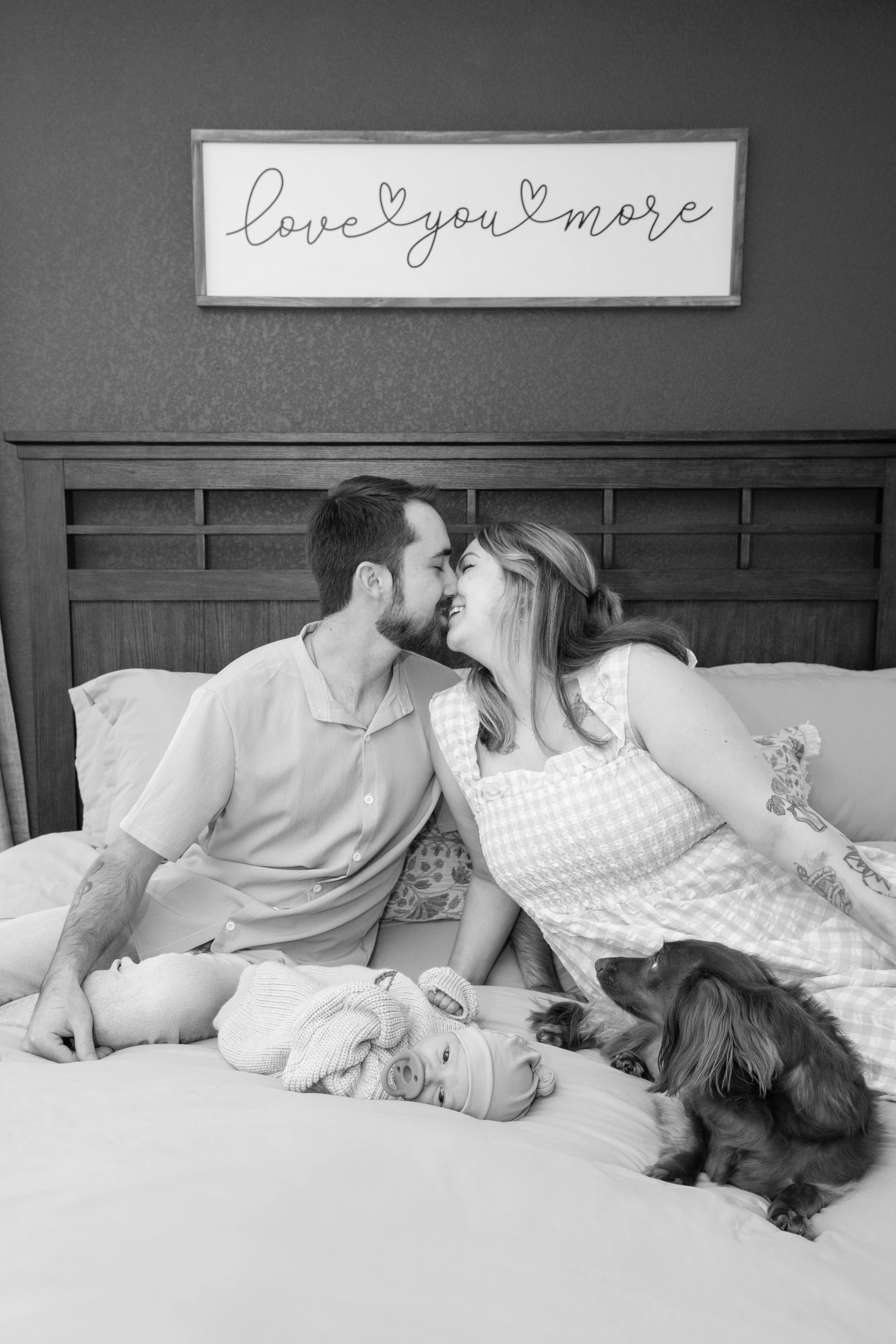A black and white photo of a couple and a baby on a bed with a dog, and a sign that says 'love you more' on the wall behind them.