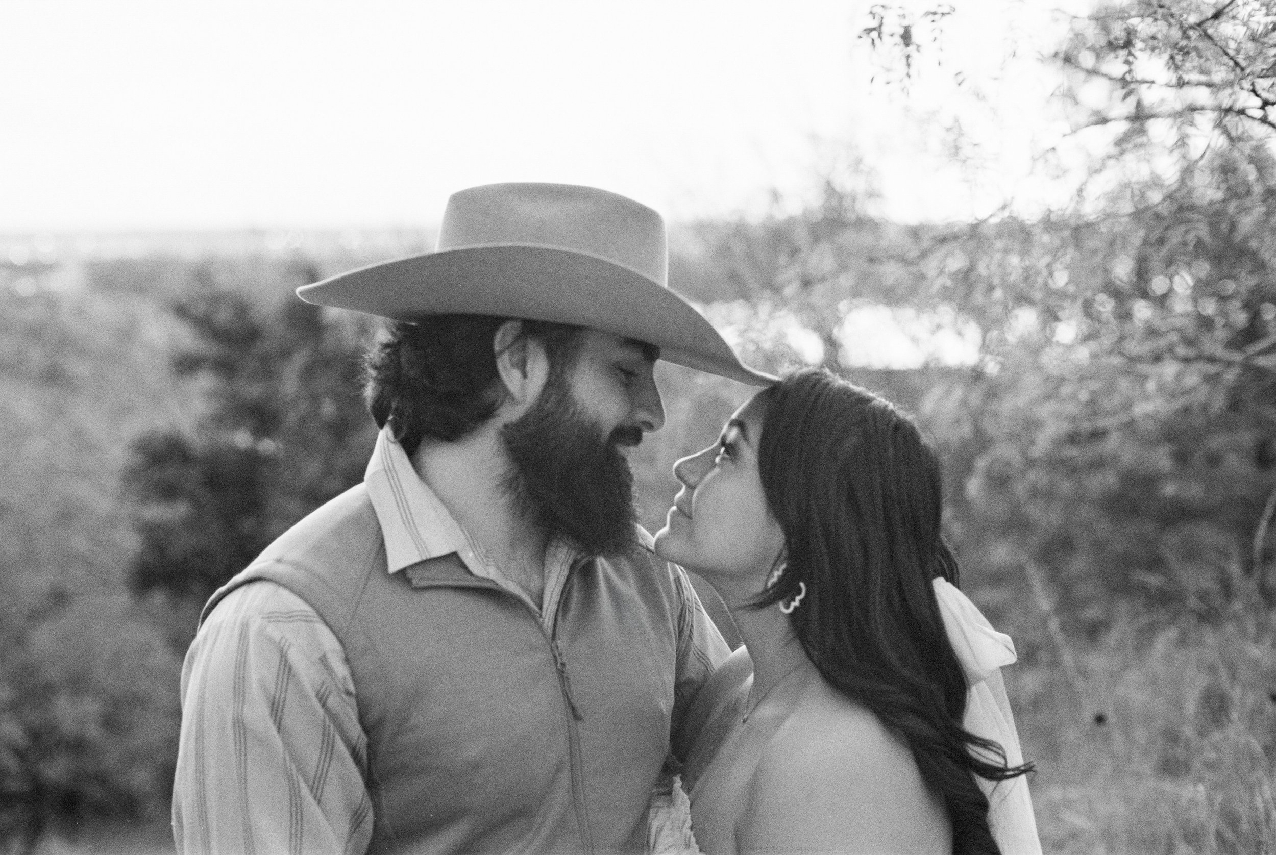 A man with a beard wearing a cowboy hat and a vest, and a woman with long dark hair and earrings, gazing at each other lovingly outdoors in a natural setting.