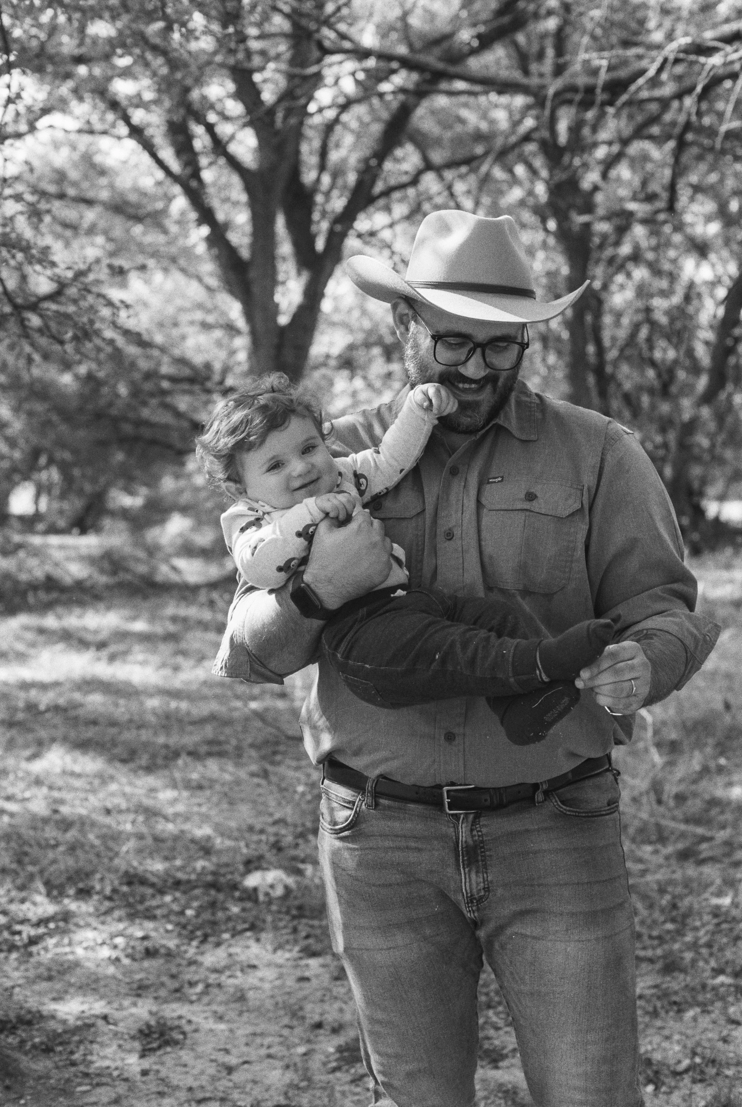 A man wearing a cowboy hat and glasses holding a smiling toddler in an outdoor park with trees in the background.