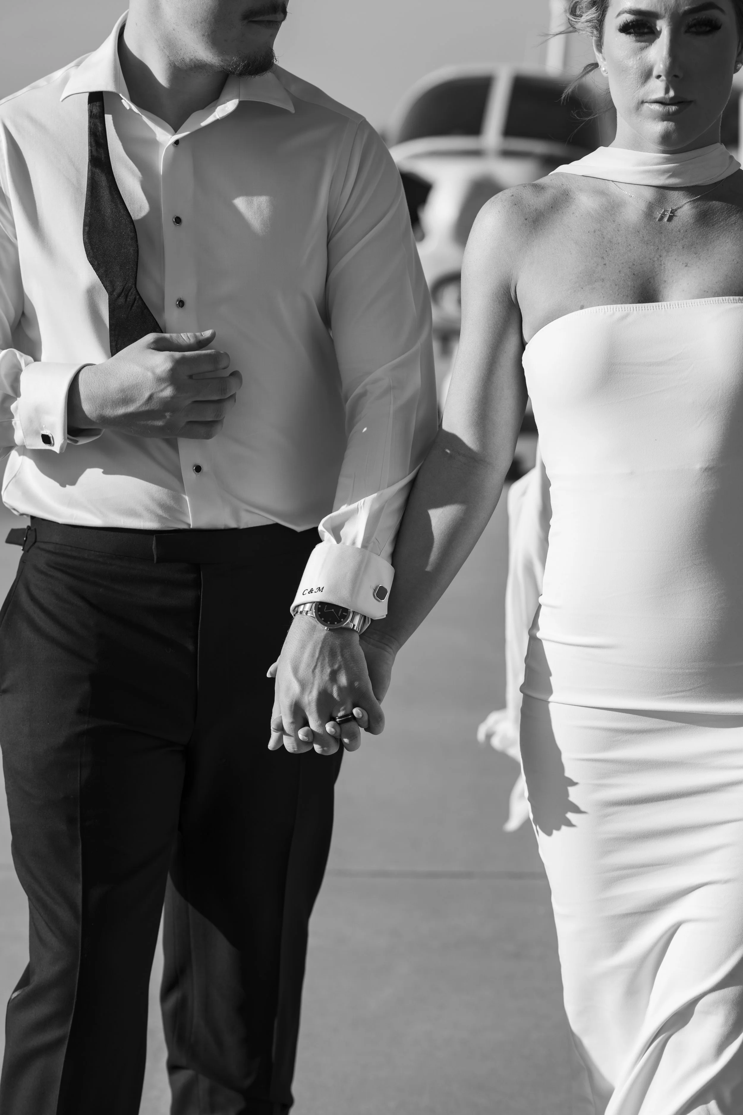 A black-and-white photo of a couple holding hands, with the man in a white dress shirt and black trousers, and the woman in a strapless white dress, possibly at a wedding or special event.