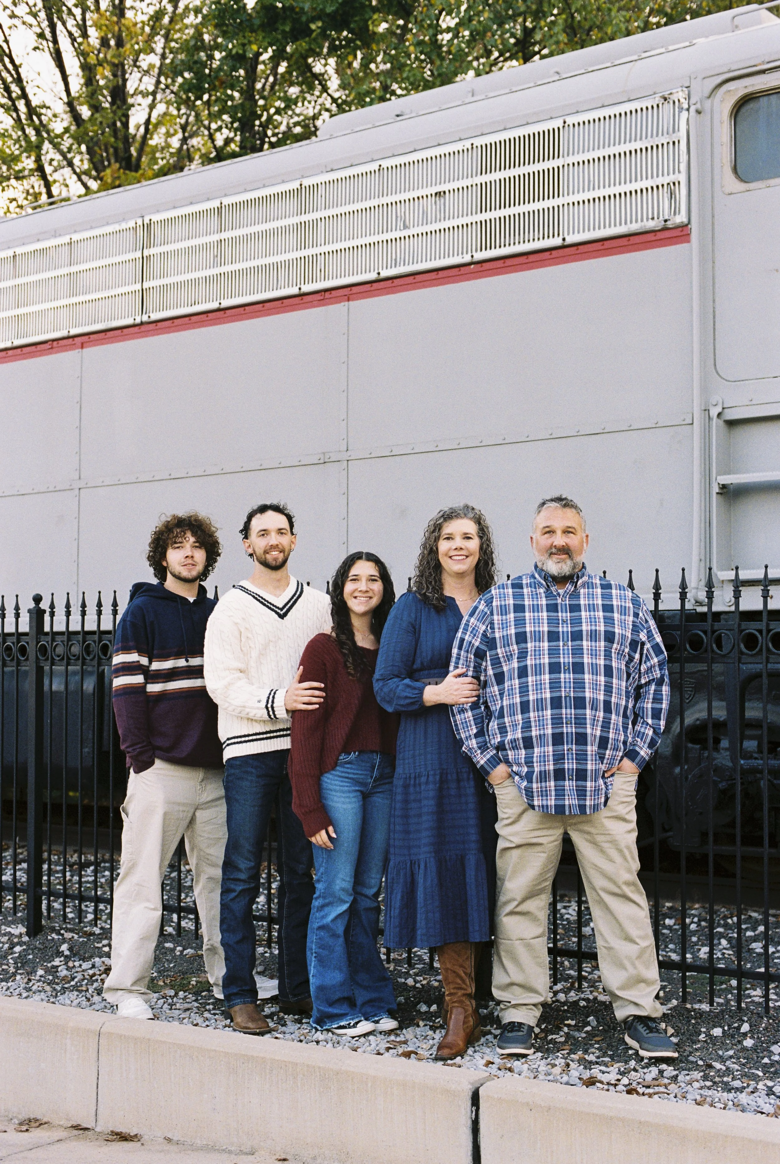 Group of five people standing in front of a fence and a large vehicle, possibly a bus or train, outdoors during daytime.