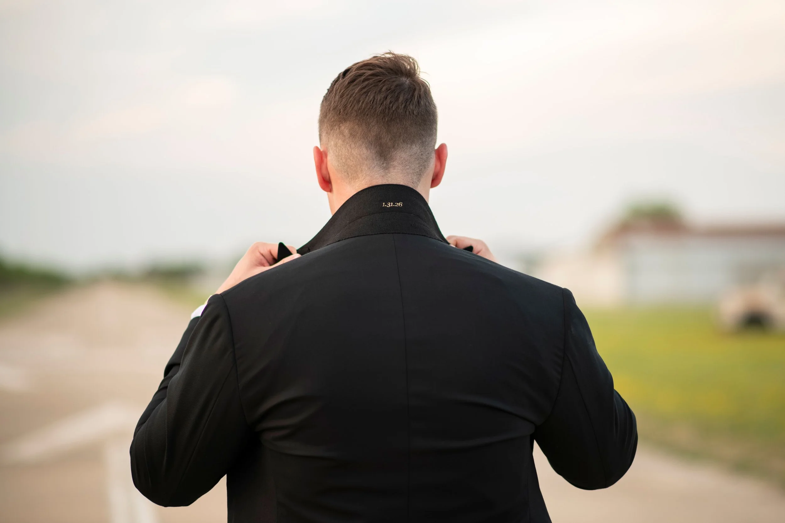 A man seen from behind, wearing a black suit and adjusting the collar, in an outdoor setting with a blurred background of open sky and distant landscape.