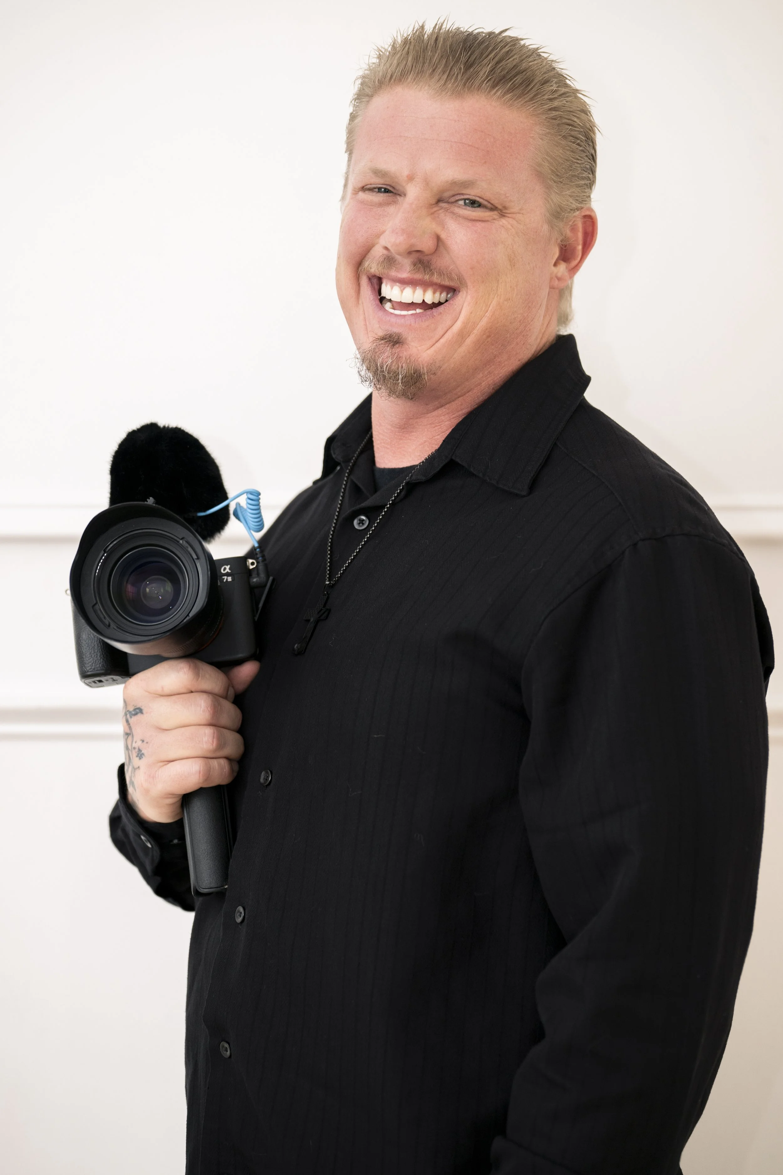 A middle-aged man with blond hair, a short beard, and mustache, smiling and holding a camera with a microphone attached, dressed in a black shirt, standing against a white background.