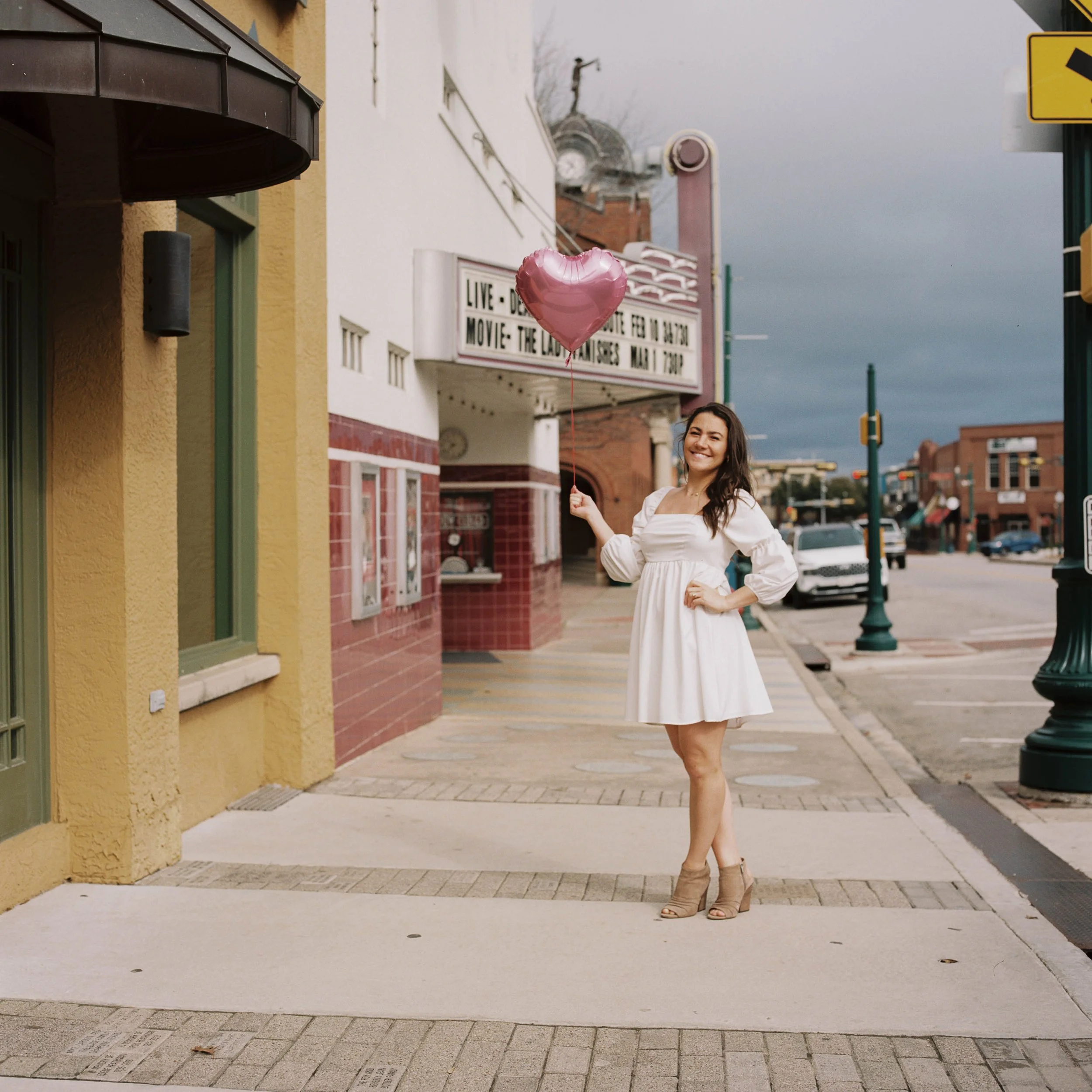 A woman in a white dress holding a pink heart-shaped balloon on a city sidewalk with buildings, cars, and a movie theater marquee in the background.