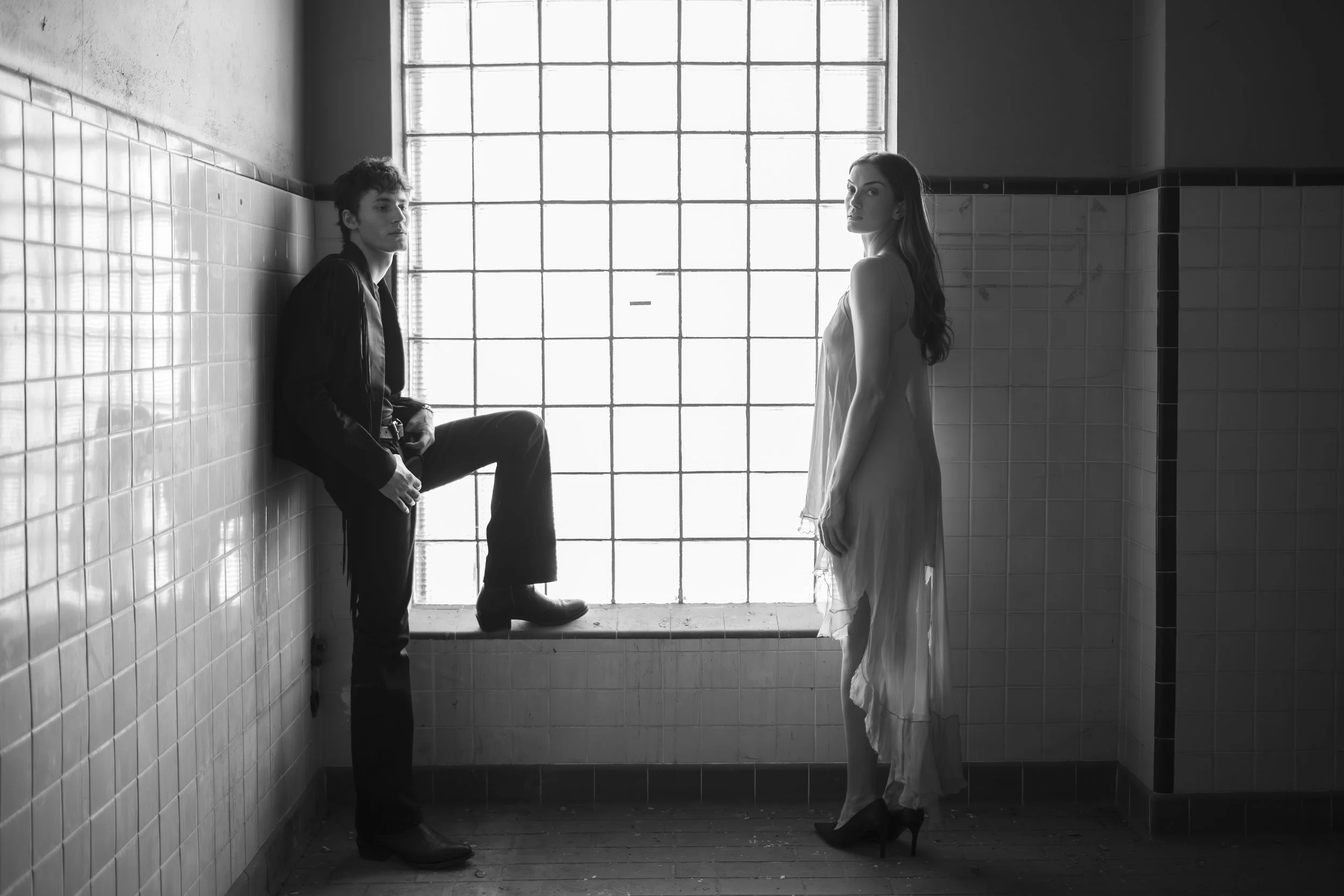 A black-and-white photograph of a man and woman standing in a tiled room near a large glass window. The man is sitting on a ledge, wearing dark clothing, and the woman is standing, wearing a light dress and high heels. They are facing each other, wit