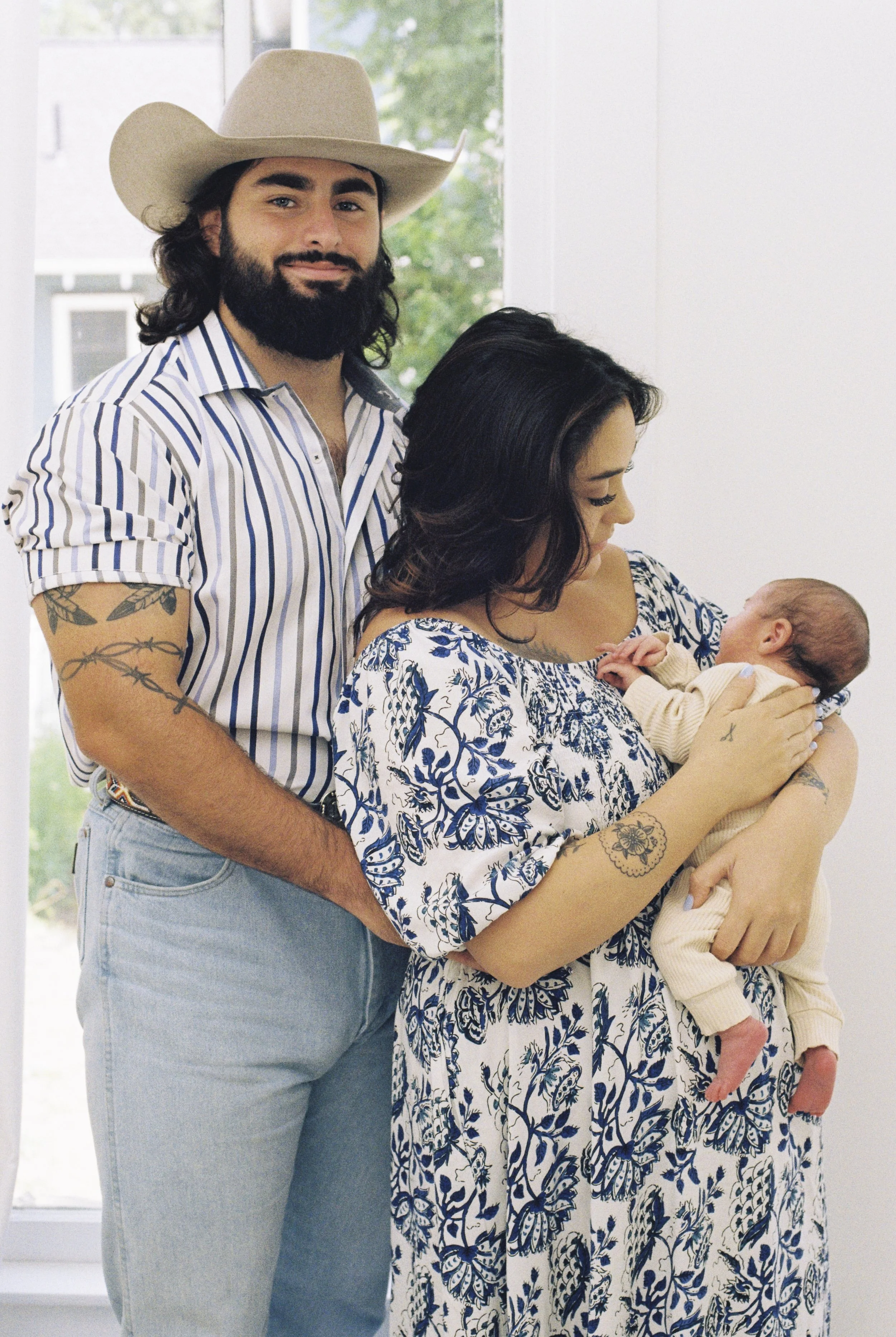 A man wearing a cowboy hat and striped shirt standing behind a woman holding a newborn baby, with a window and greenery outside in the background.