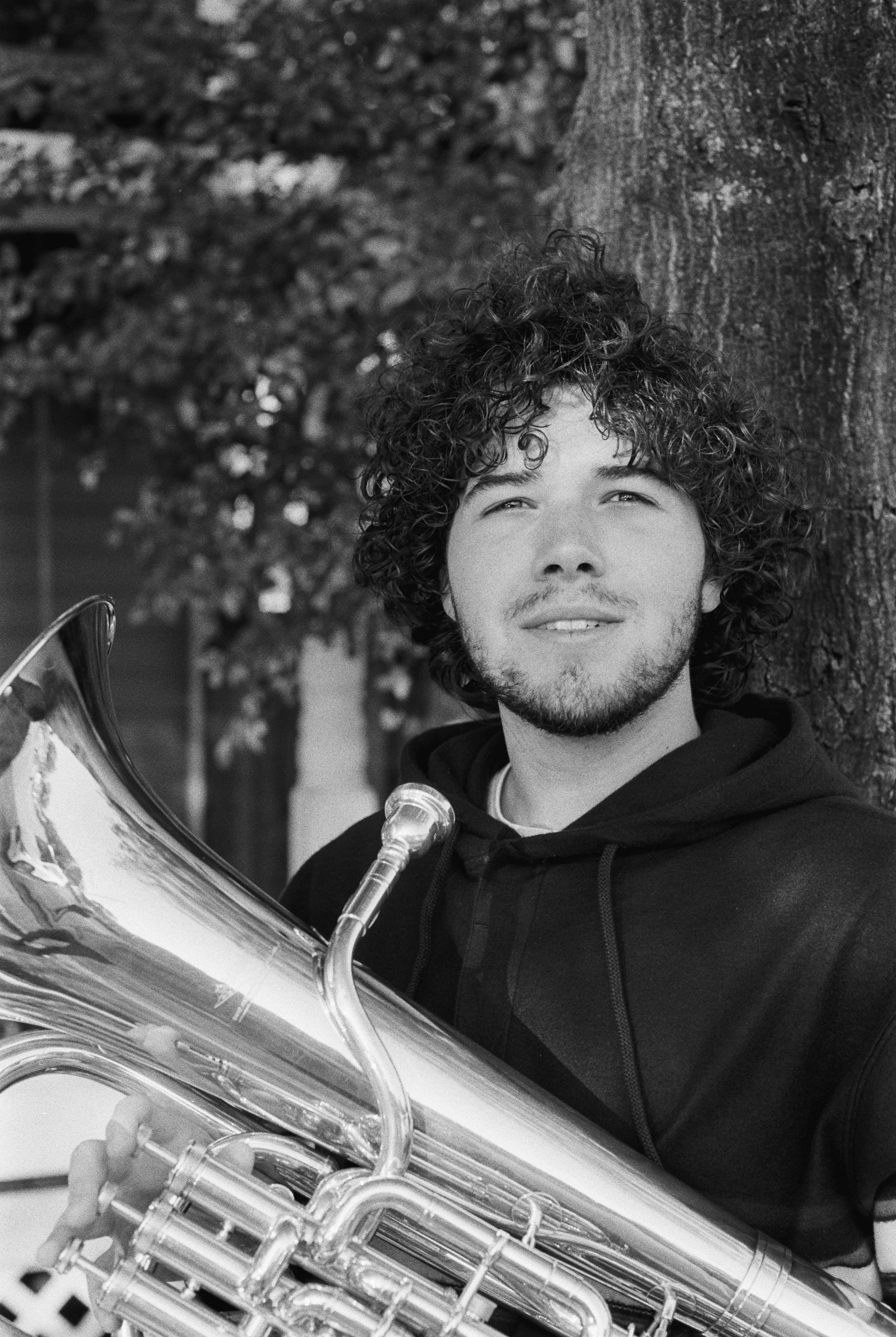 Black and white photo of a young man with curly hair, a beard, and light eyes, leaning against a tree and holding a shiny saxophone.