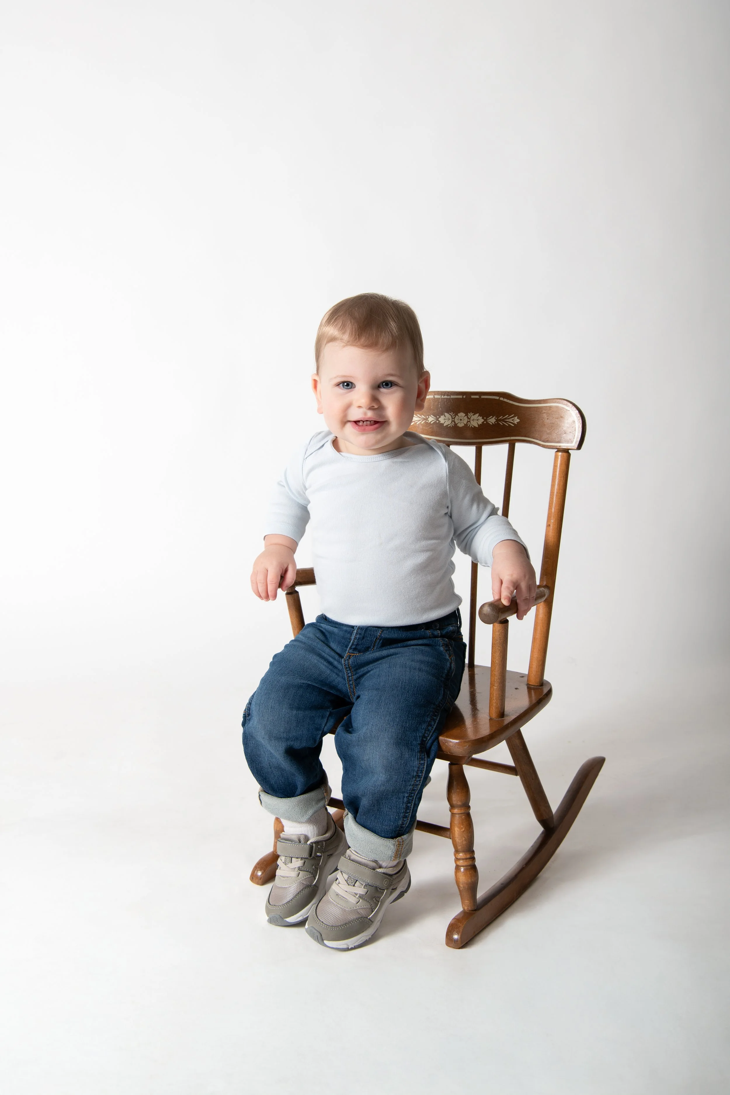 A young boy sitting on a wooden rocking chair, wearing a white long-sleeve shirt, blue jeans, and gray sneakers, smiling at the camera against a plain white background.