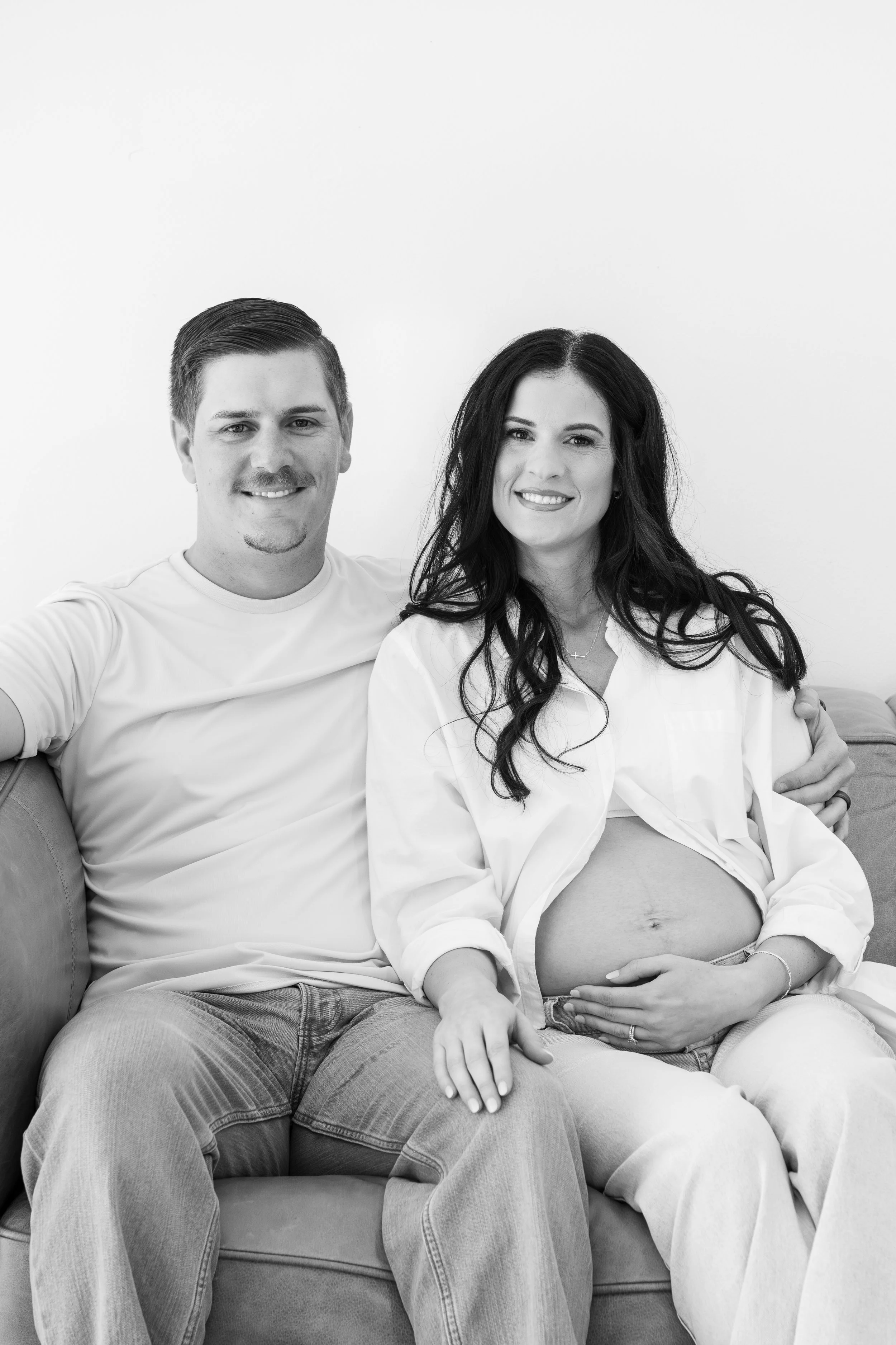 A couple sitting on a couch with the woman visibly pregnant, smiling at the camera, in a black and white photo.