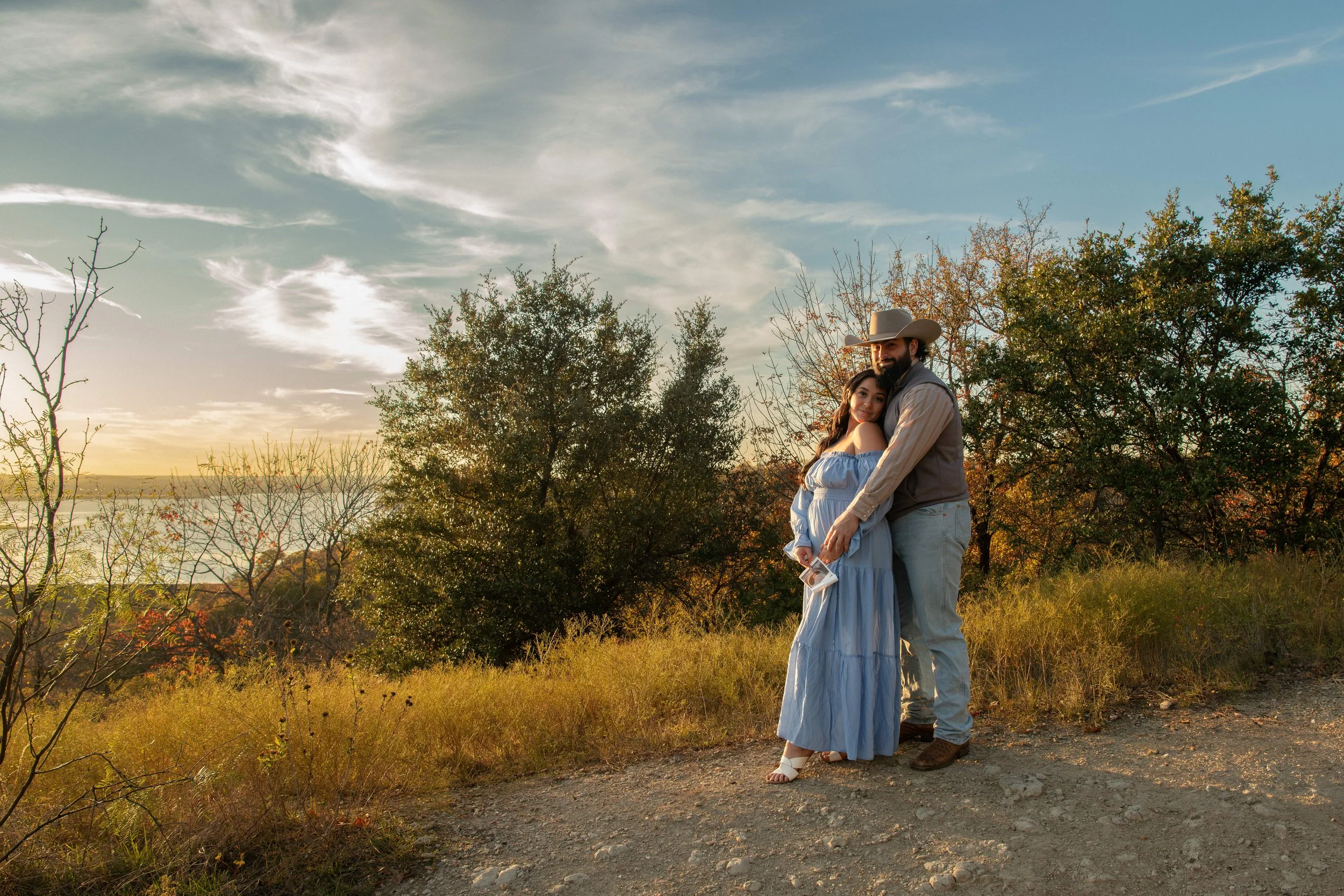 A couple standing on a dirt path outdoors during sunset, hugging each other. The woman is wearing a light blue off-shoulder dress, and the man is wearing a cowboy hat, a vest, and jeans. Behind them are trees and a body of water in the distance.