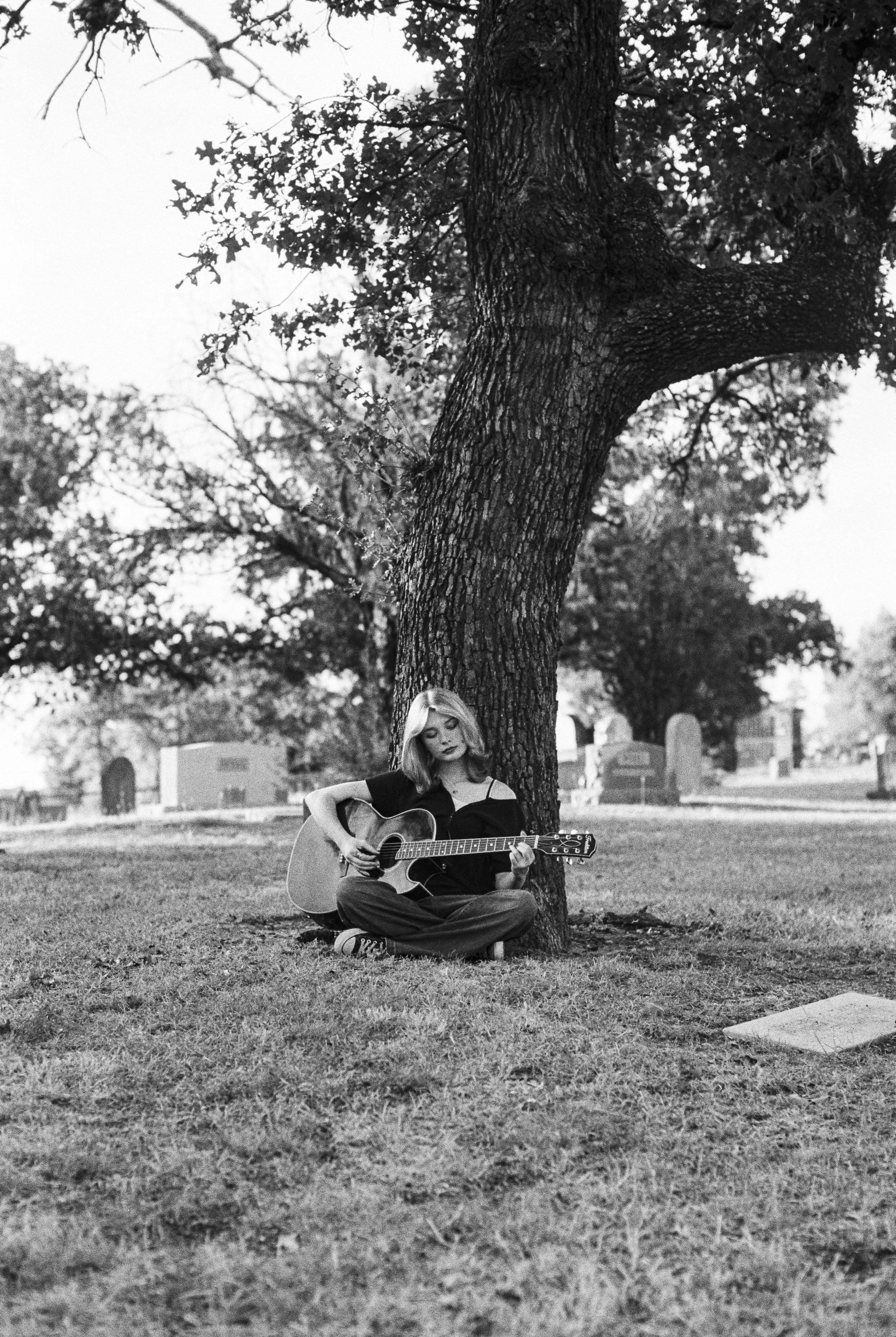 A young woman with shoulder-length hair, dressed in dark clothing, sits cross-legged on the grass and plays an acoustic guitar under a large tree in a graveyard, with tombstones visible in the background. Black and white photo.