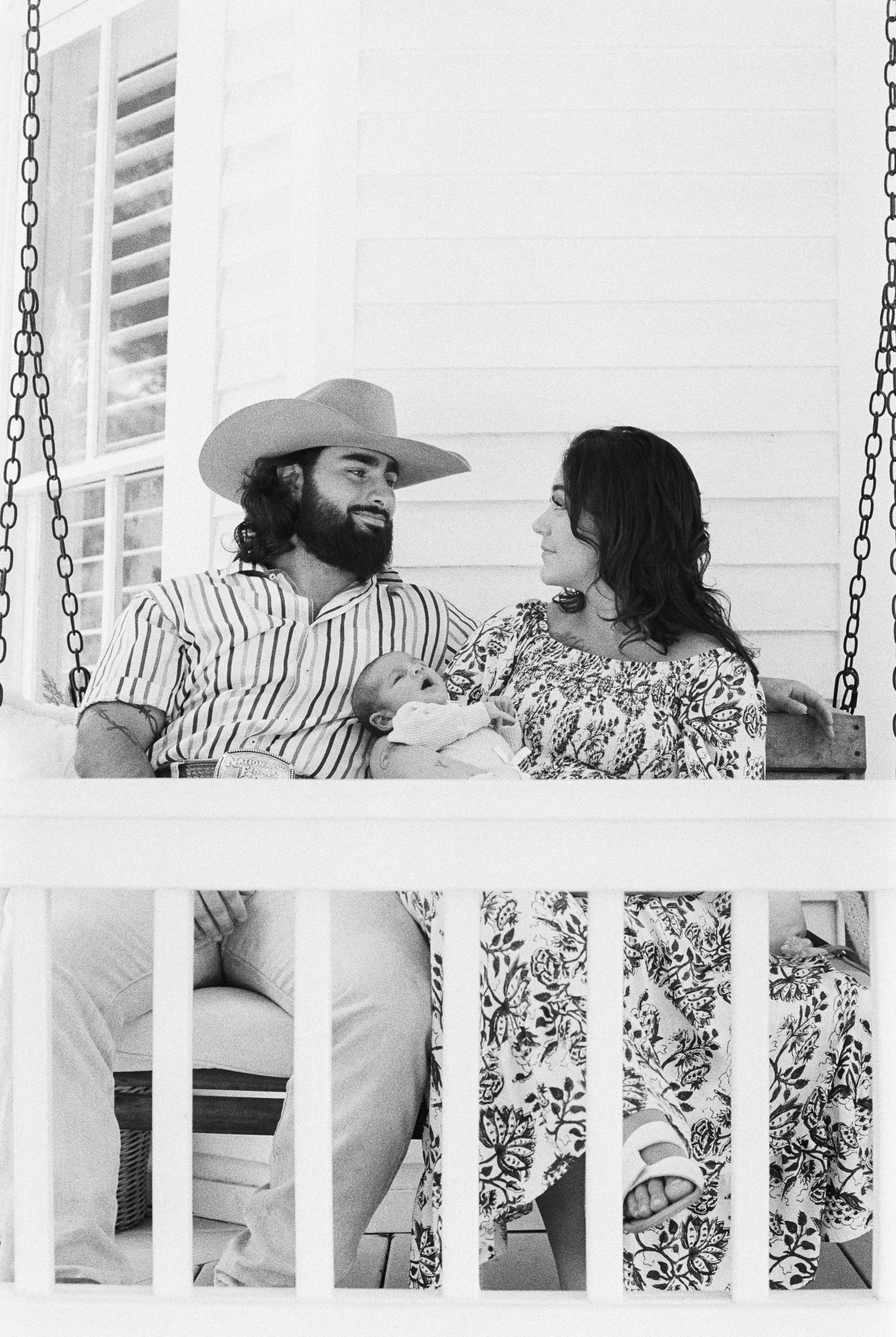 A couple sitting on a porch swing holding a baby, with the woman looking at the man and the man looking at the woman, in black and white.