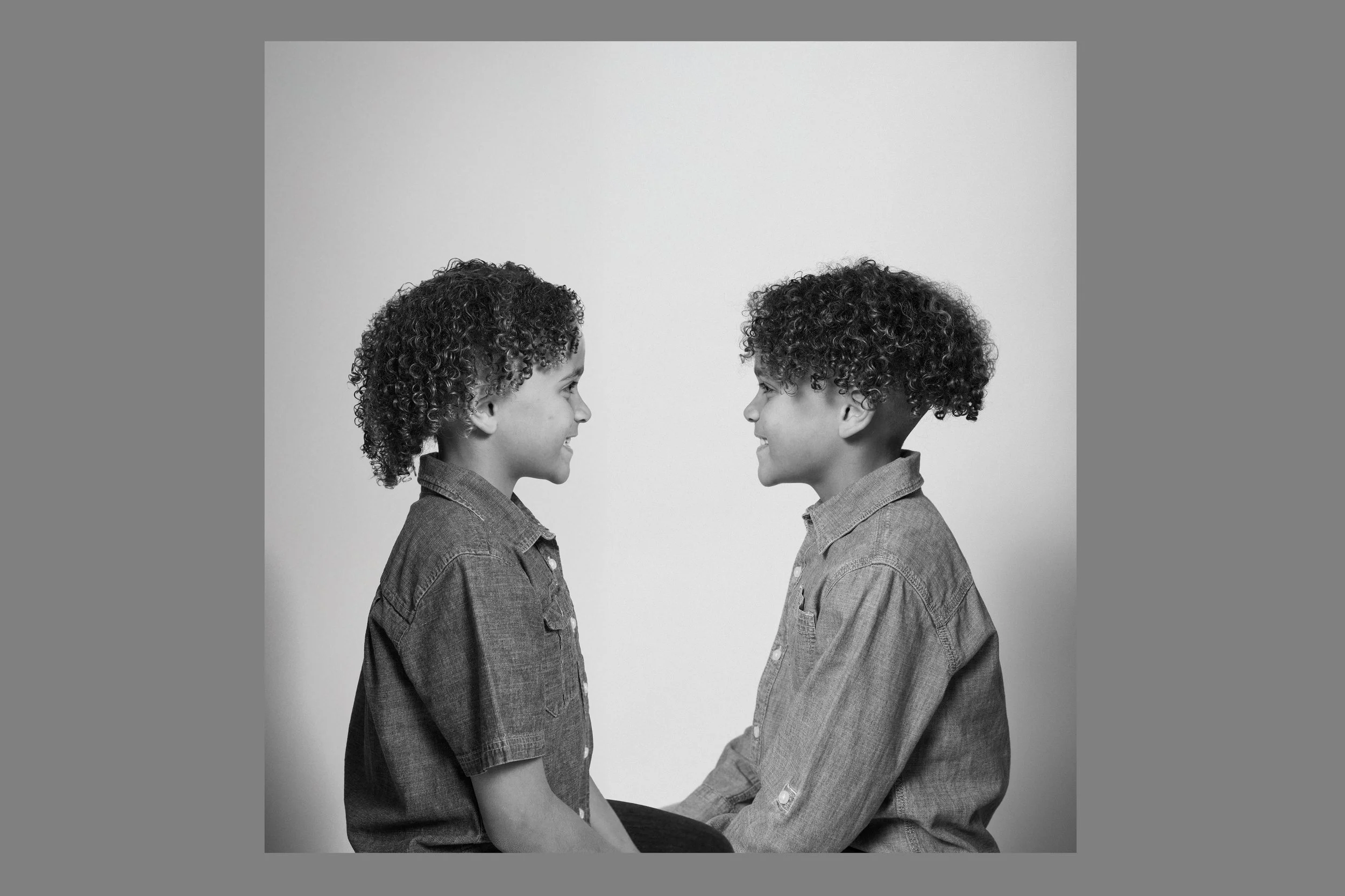 Two children with curly hair, one girl and one boy, facing each other, smiling, wearing denim shirts, sitting on the floor against a plain background.