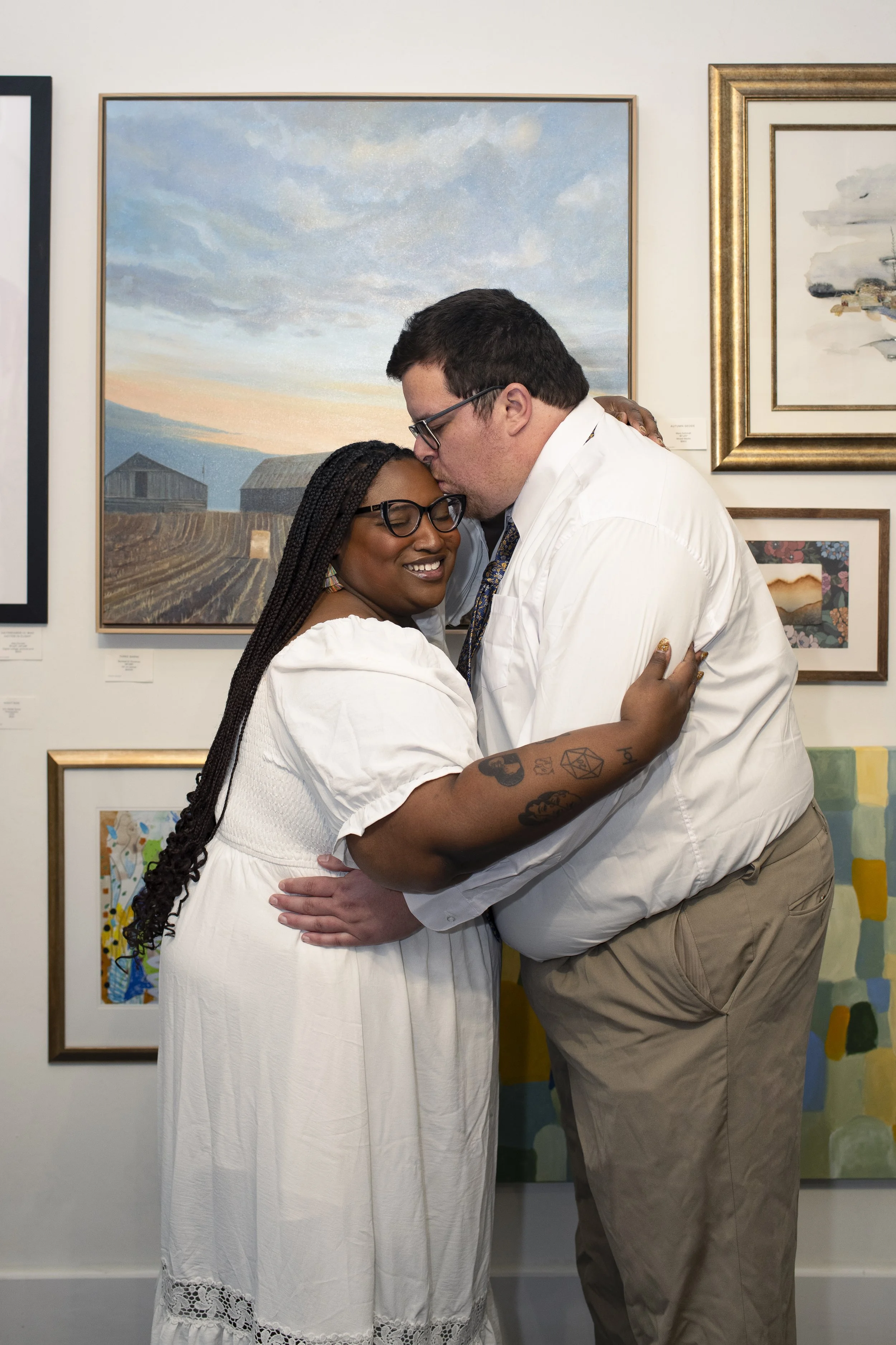 A couple embracing at an art gallery, with the man kissing the woman's forehead while she smiles with eyes closed. They are both wearing white tops, and the background features various framed paintings on the wall.